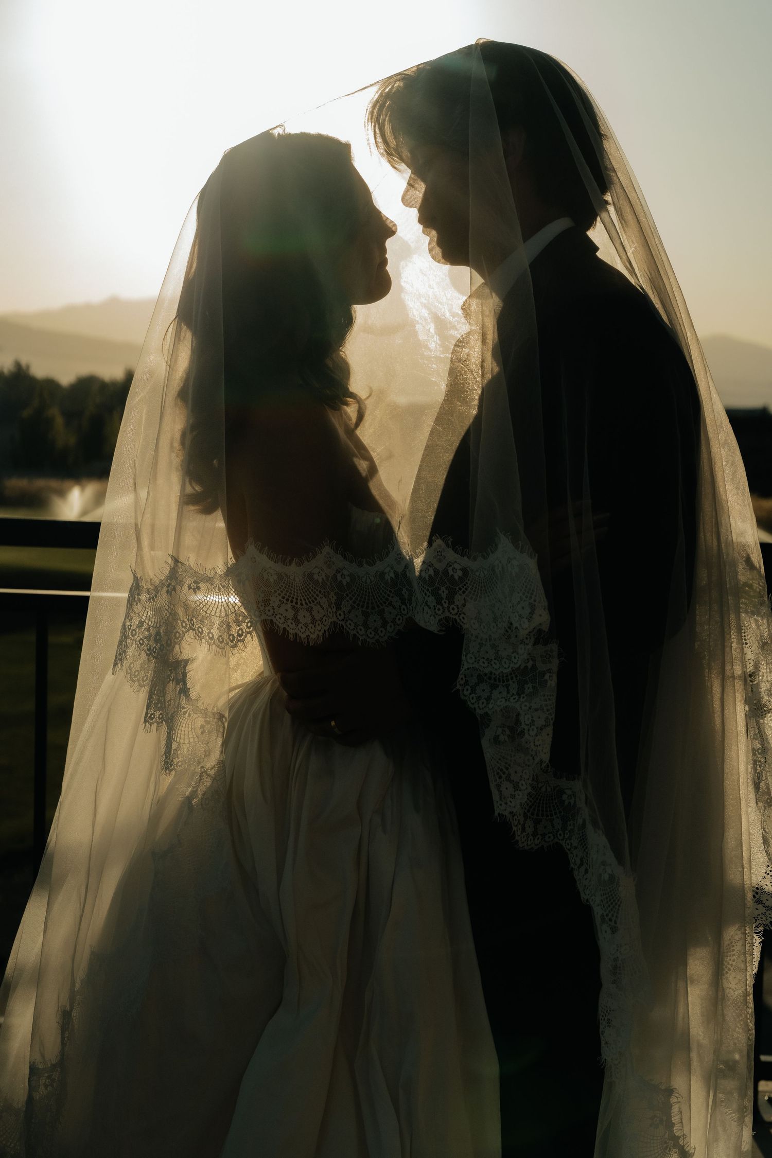 Romantic backlit silhouette of bride and groom under wedding veil at sunset with mountains in background.