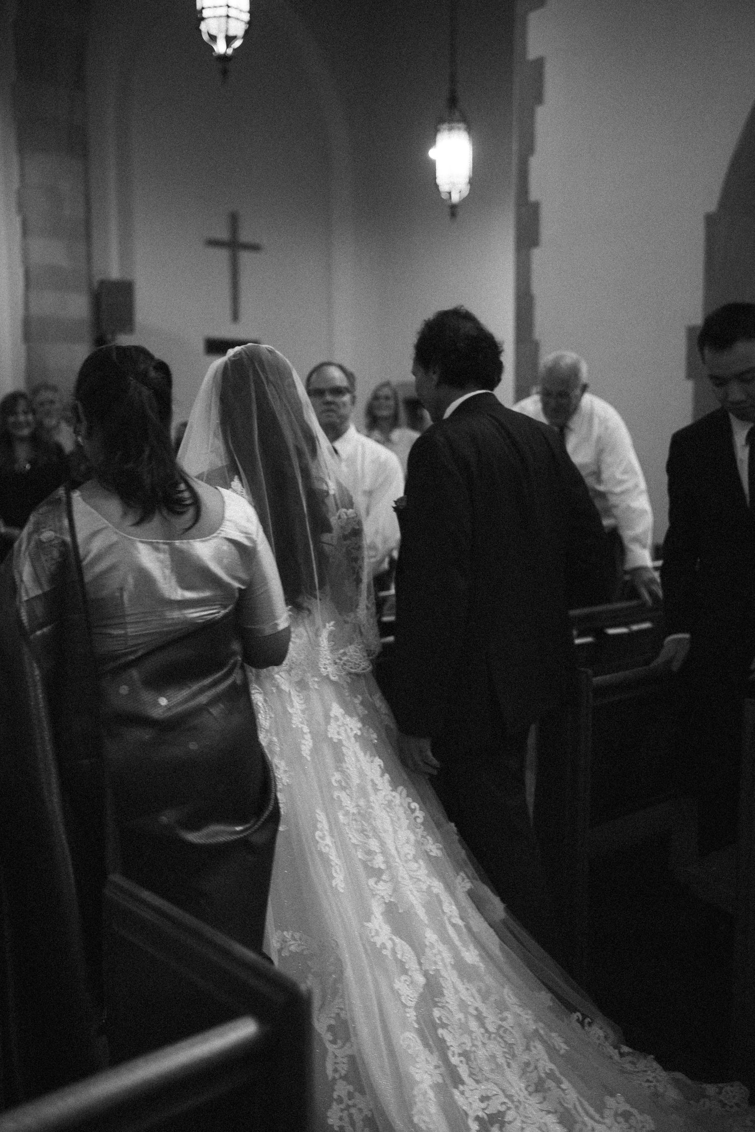 Black and white photo of a bride in lace gown walking down church aisle with wedding party toward altar cross.
