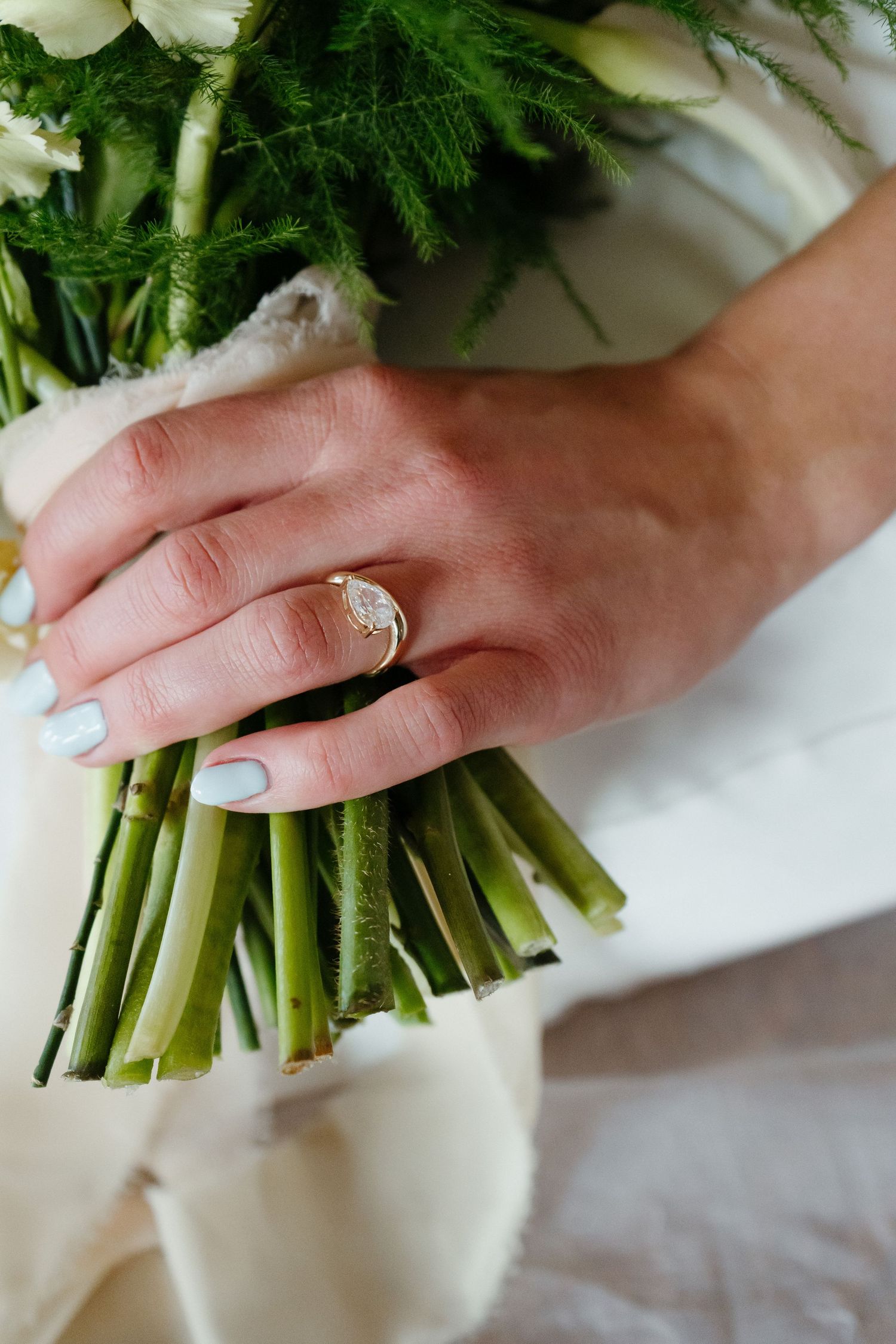 Close-up of brides hand with oval diamond ring holding green stemmed wedding bouquet.