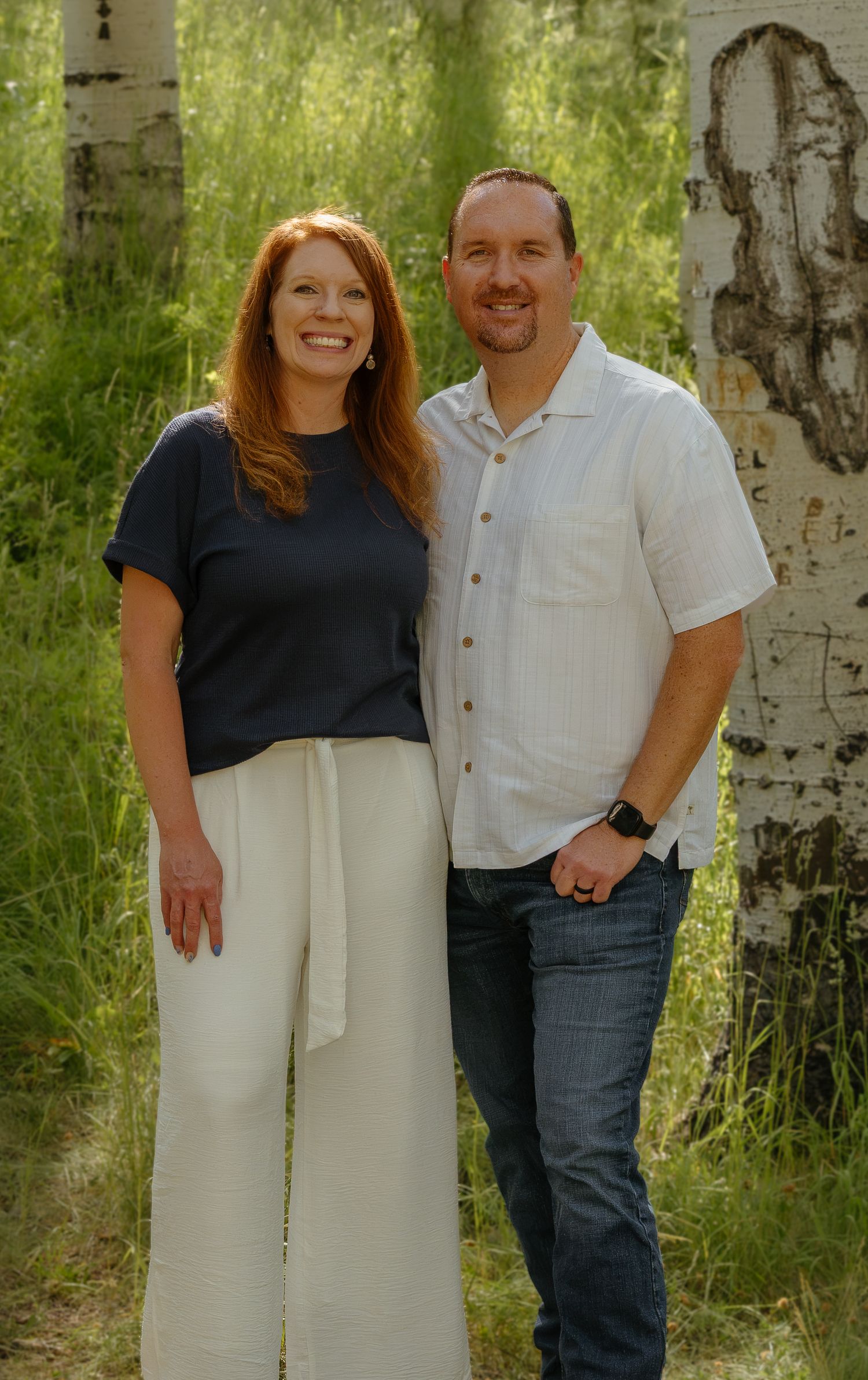 A smiling couple poses outdoors among aspen trees in a lush green forest setting.