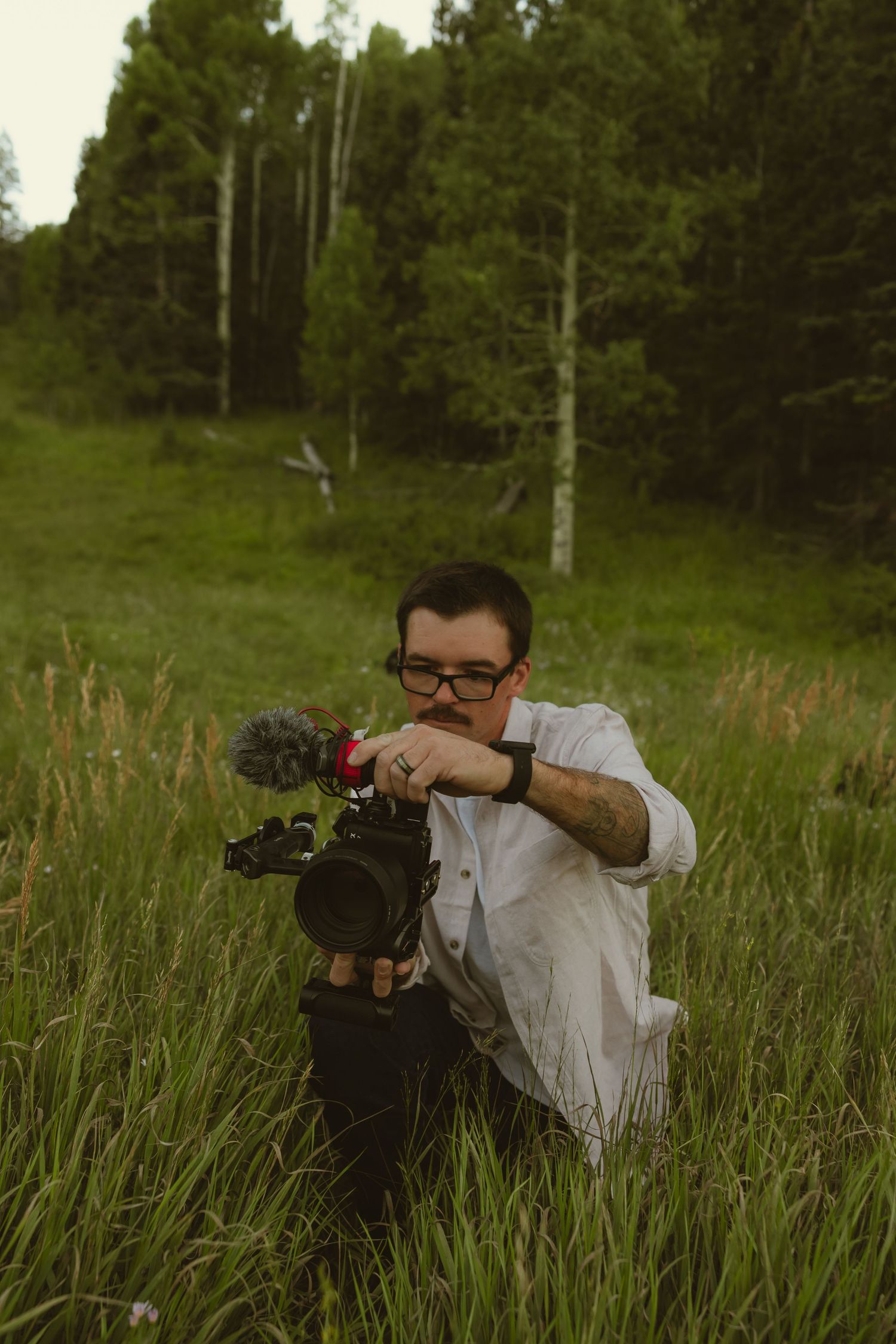 Photographer crouching in tall grass holding a camera with microphone attachment in a lush green forest meadow.