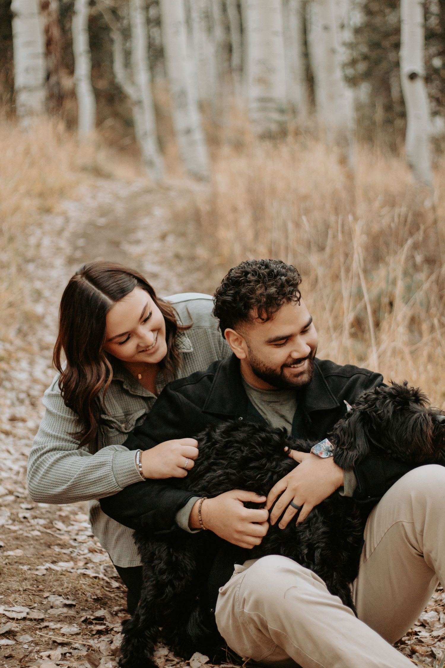 Couple smiling and cuddling with a black dog on a forest trail surrounded by autumn foliage.