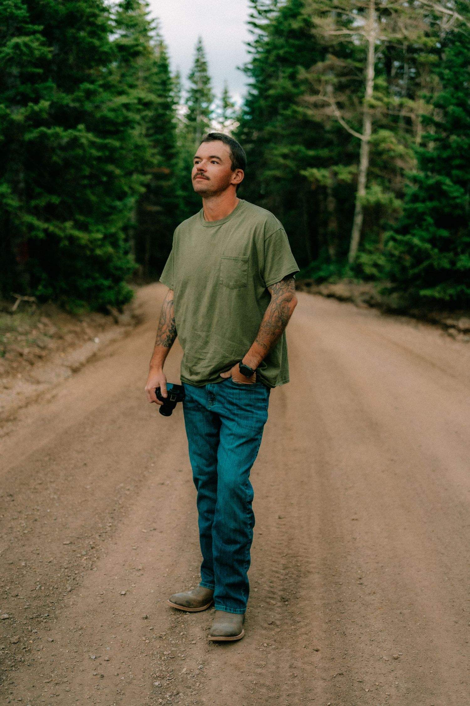 Tattooed man in green t-shirt and teal jeans walking on a dirt forest road surrounded by tall pine trees.