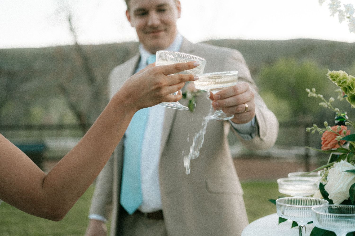 Wedding couple toasting with champagne glasses near floral centerpiece at outdoor reception.