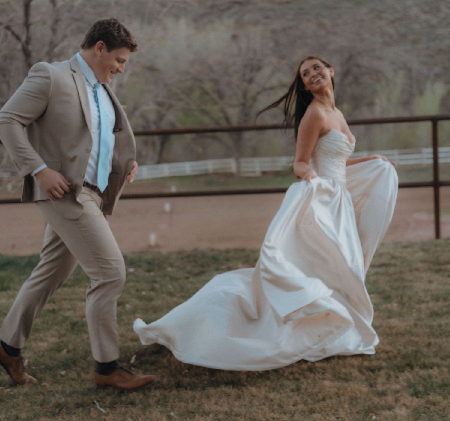 Joyful bride twirling in flowing white gown while groom chases her at outdoor wedding venue.