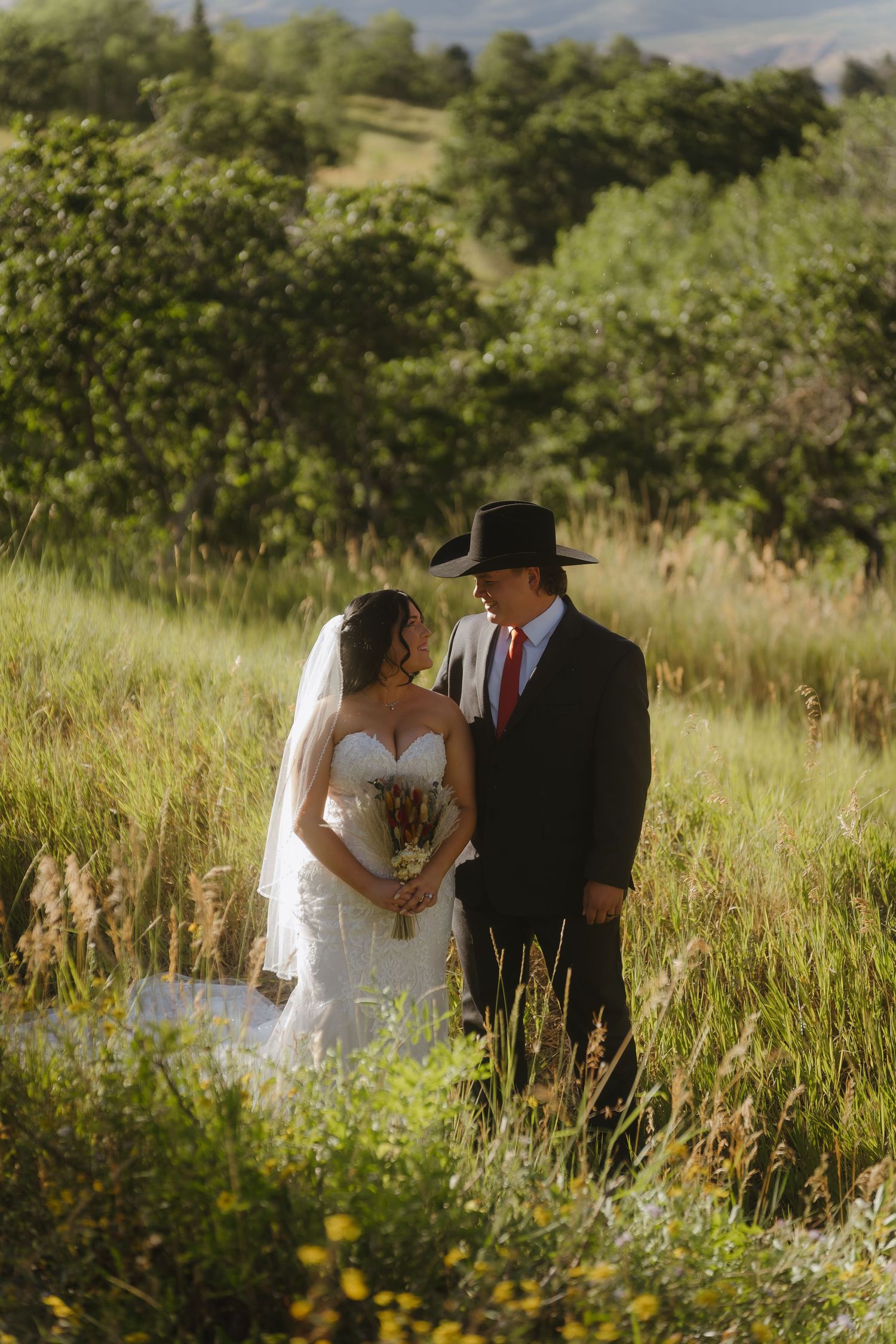 Bride and groom in cowboy hat share romantic moment in lush green meadow during outdoor western wedding ceremony.