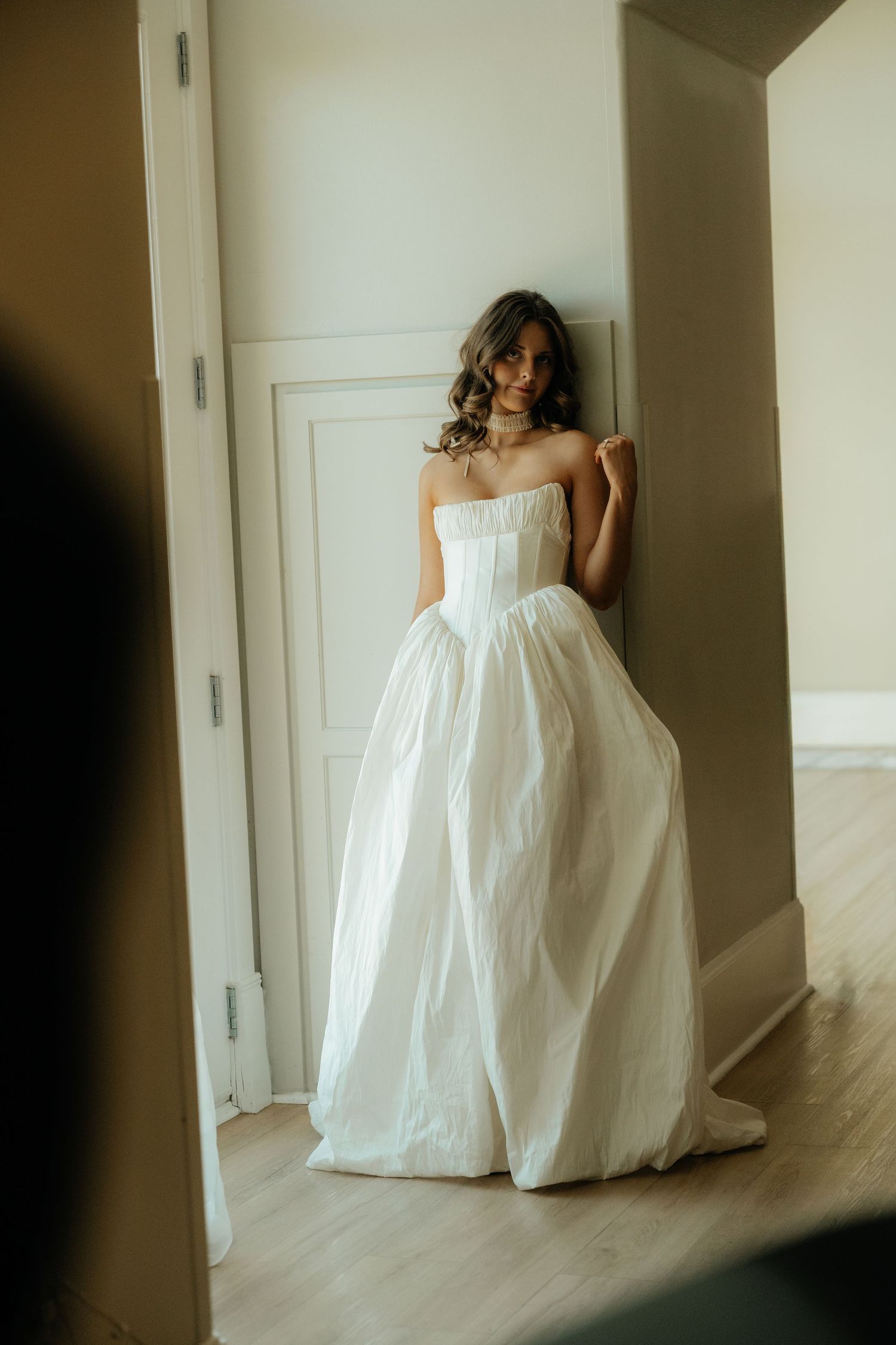 A bride in a strapless white ballgown leans against a door frame, smiling softly in an elegant indoor setting.