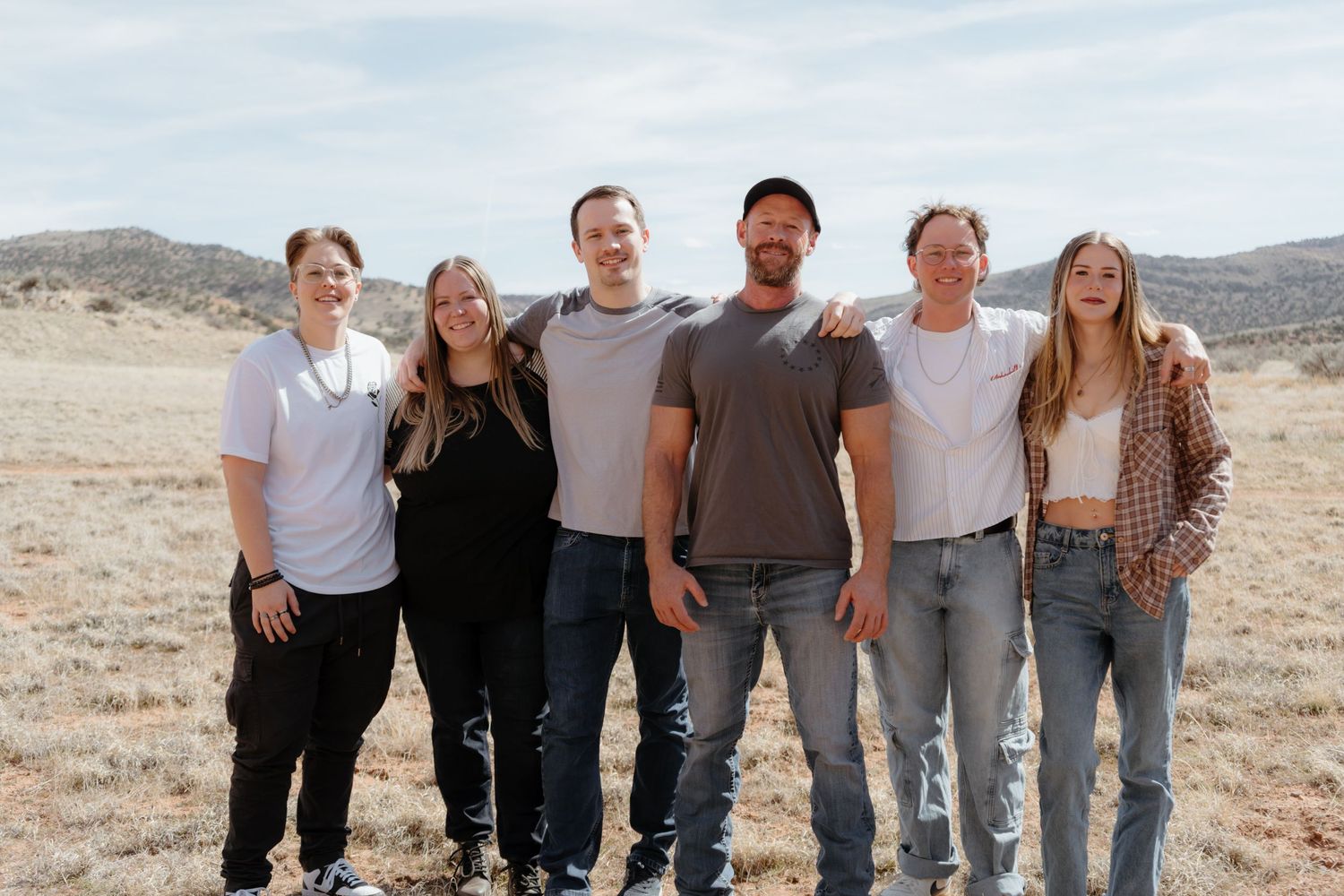 Six people smiling and posing together outdoors in a dry desert landscape with rolling hills in the background.