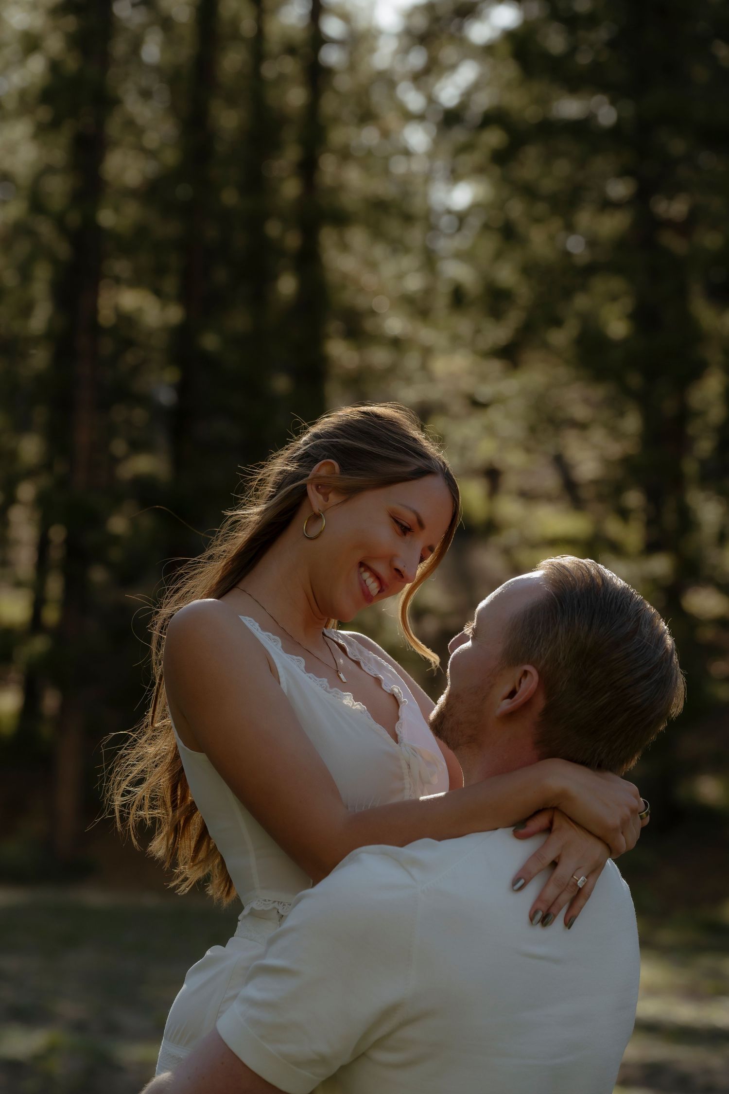 A man lifts a smiling woman in a sunlit forest, sharing a joyful romantic moment among tall pine trees.