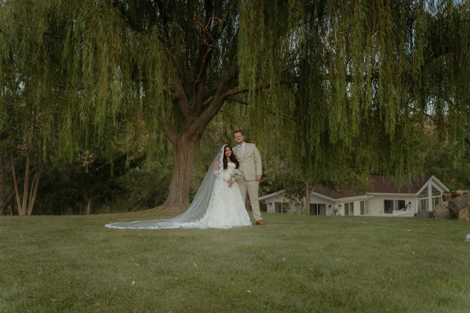 Bride and groom pose under a weeping willow tree on lush green lawn with a white farmhouse in the background.