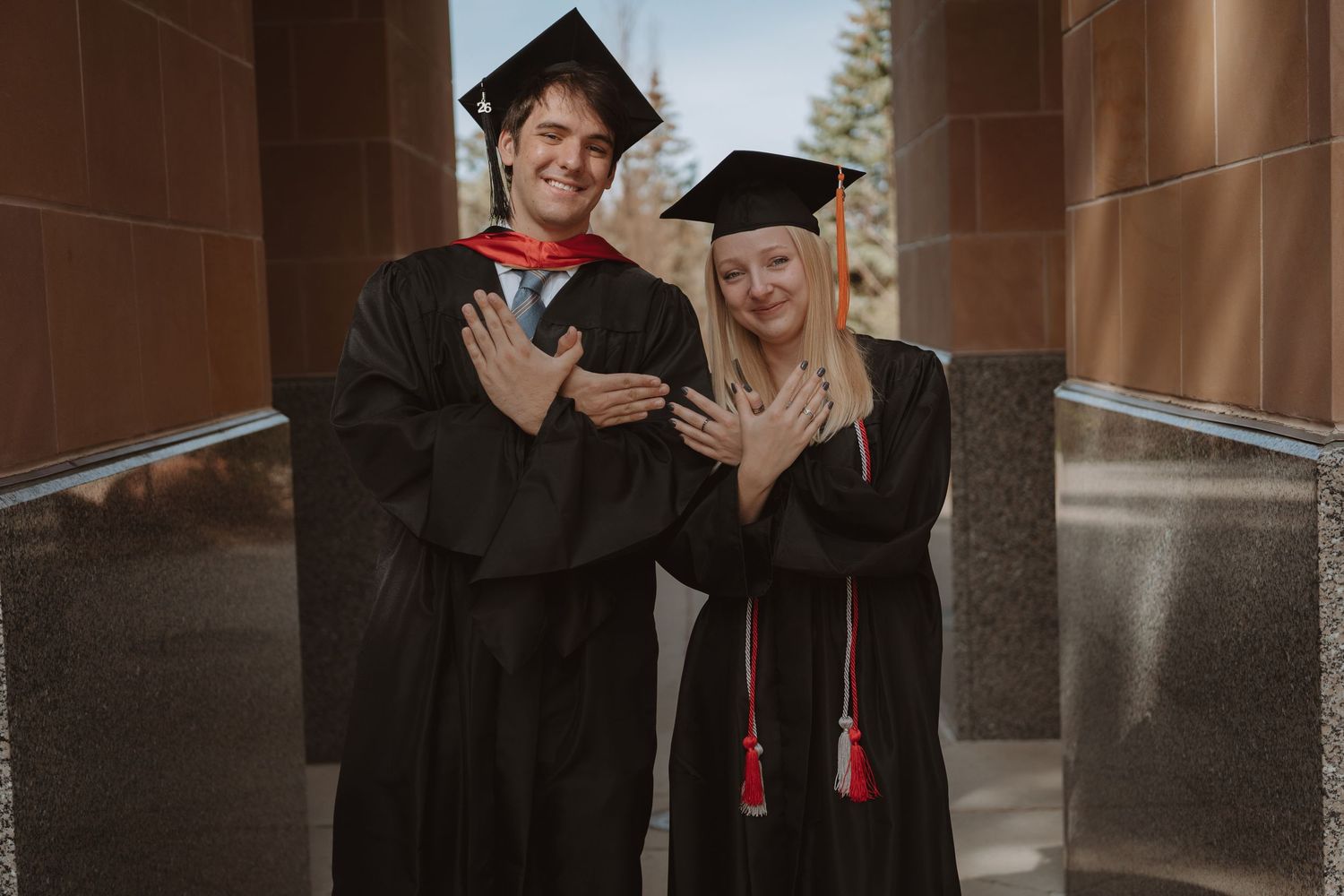 Two graduates in black caps and gowns proudly display their rings while posing together at a graduation ceremony.