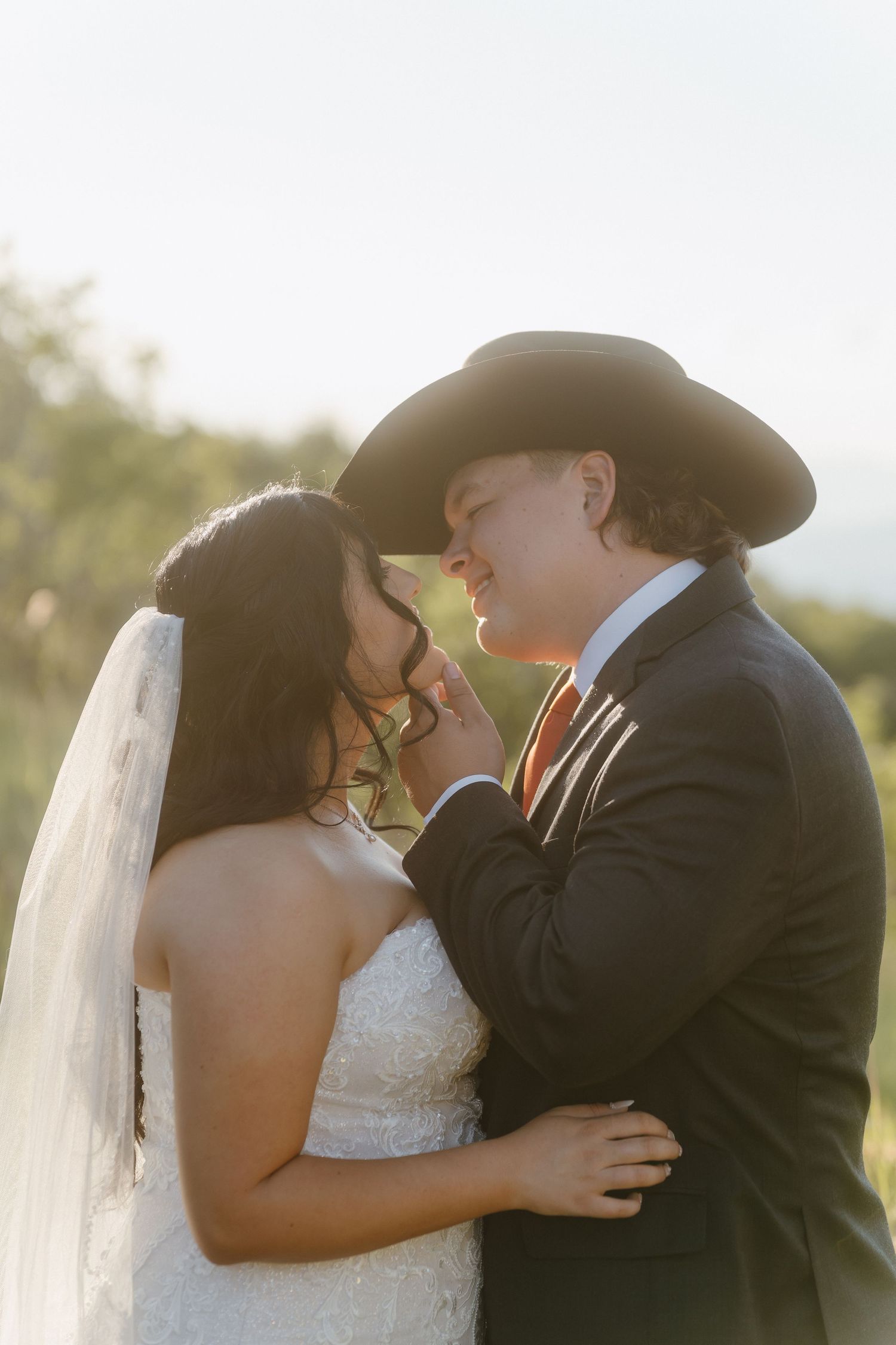 A bride and groom share a romantic moment outdoors, with the groom wearing a black cowboy hat and striped tie.