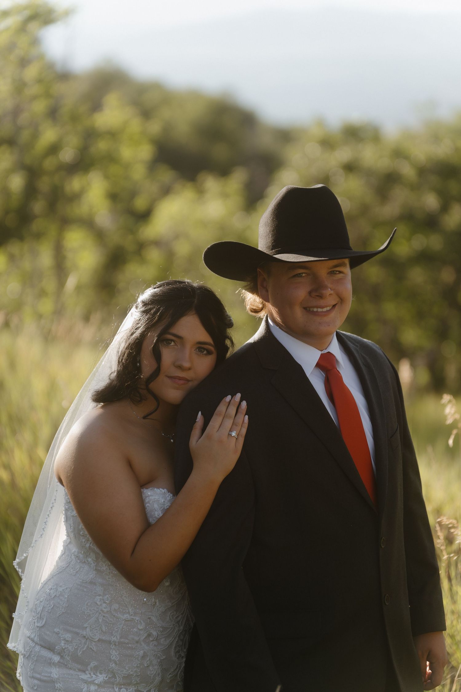 Bride in white lace gown leans on groom wearing black suit, red tie, and cowboy hat at outdoor wedding.