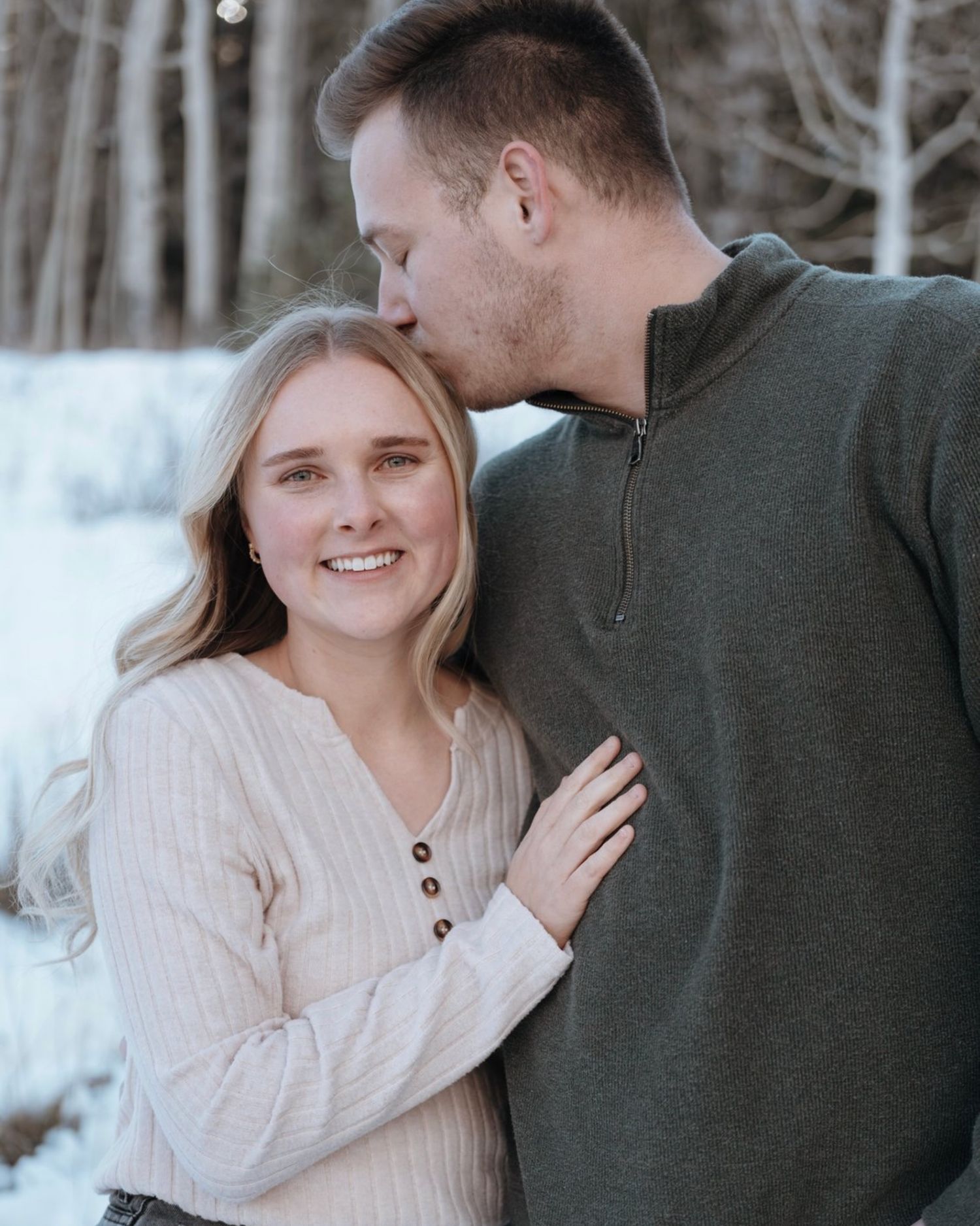 Man kissing woman on the head while she smiles in a snowy winter forest engagement photo.