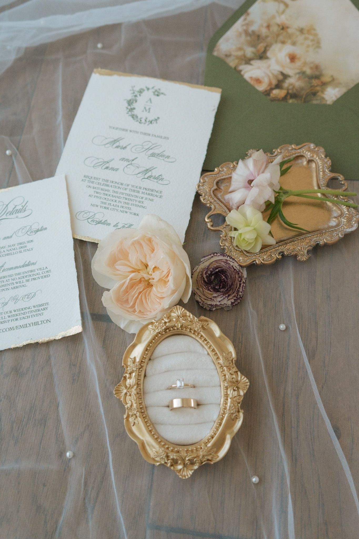 Wedding flat lay with gold rings, invitation, roses, and ornate gold frames on veil.