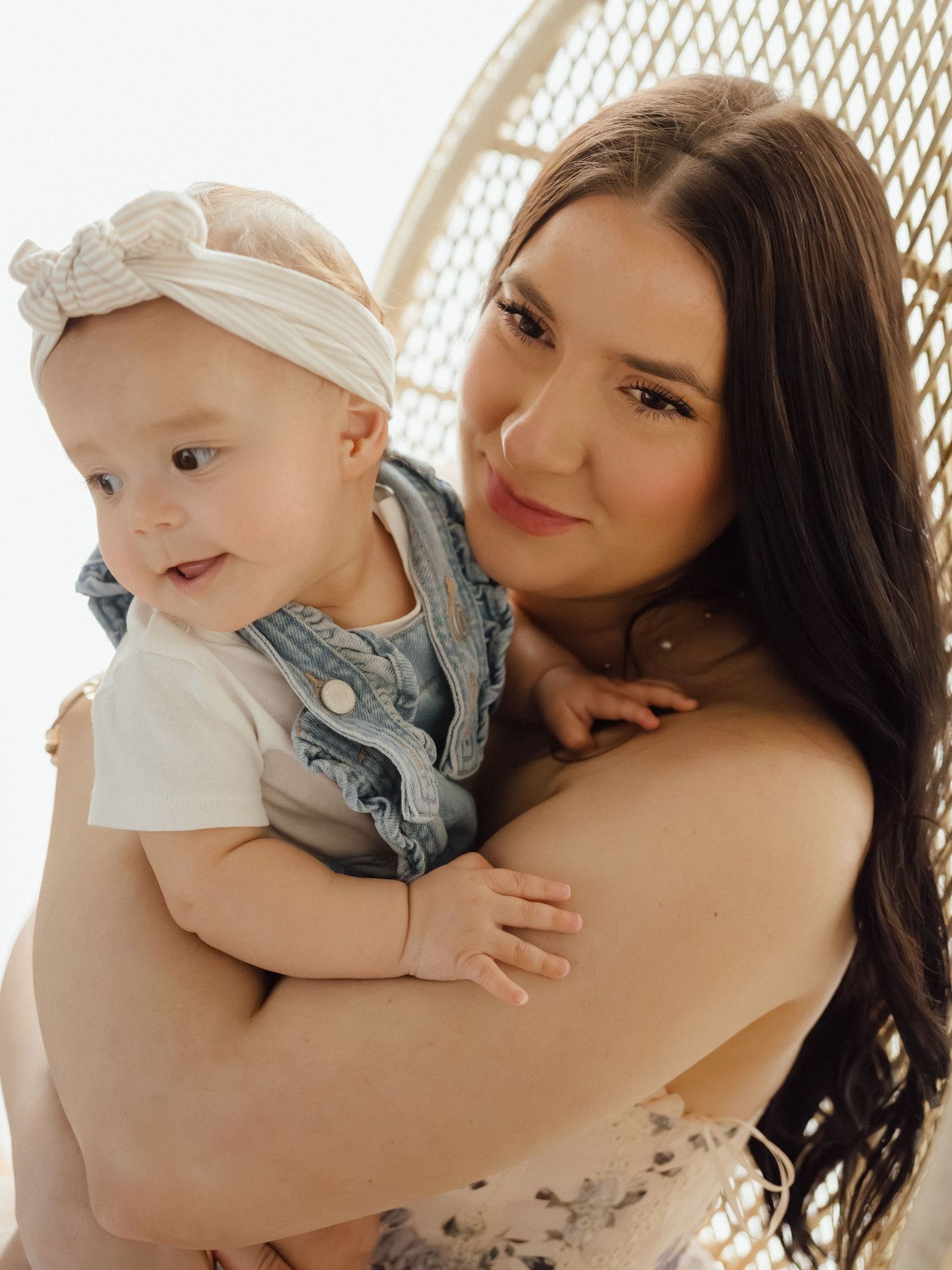 A young brunette mother smiles while holding her baby girl wearing a white headband and denim overalls.