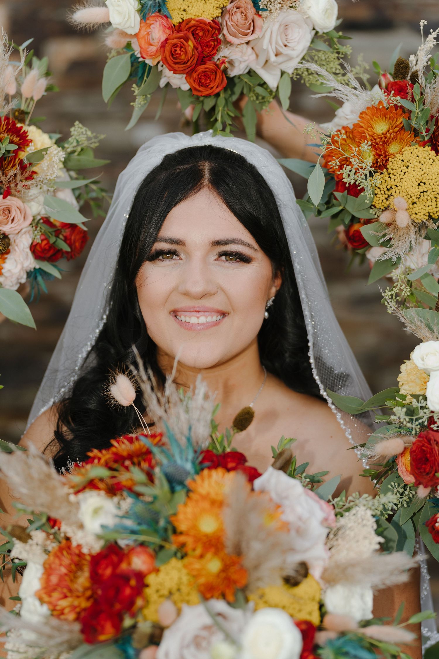 Smiling bride in white veil surrounded by vibrant fall wedding bouquets featuring orange, red, and yellow flowers.