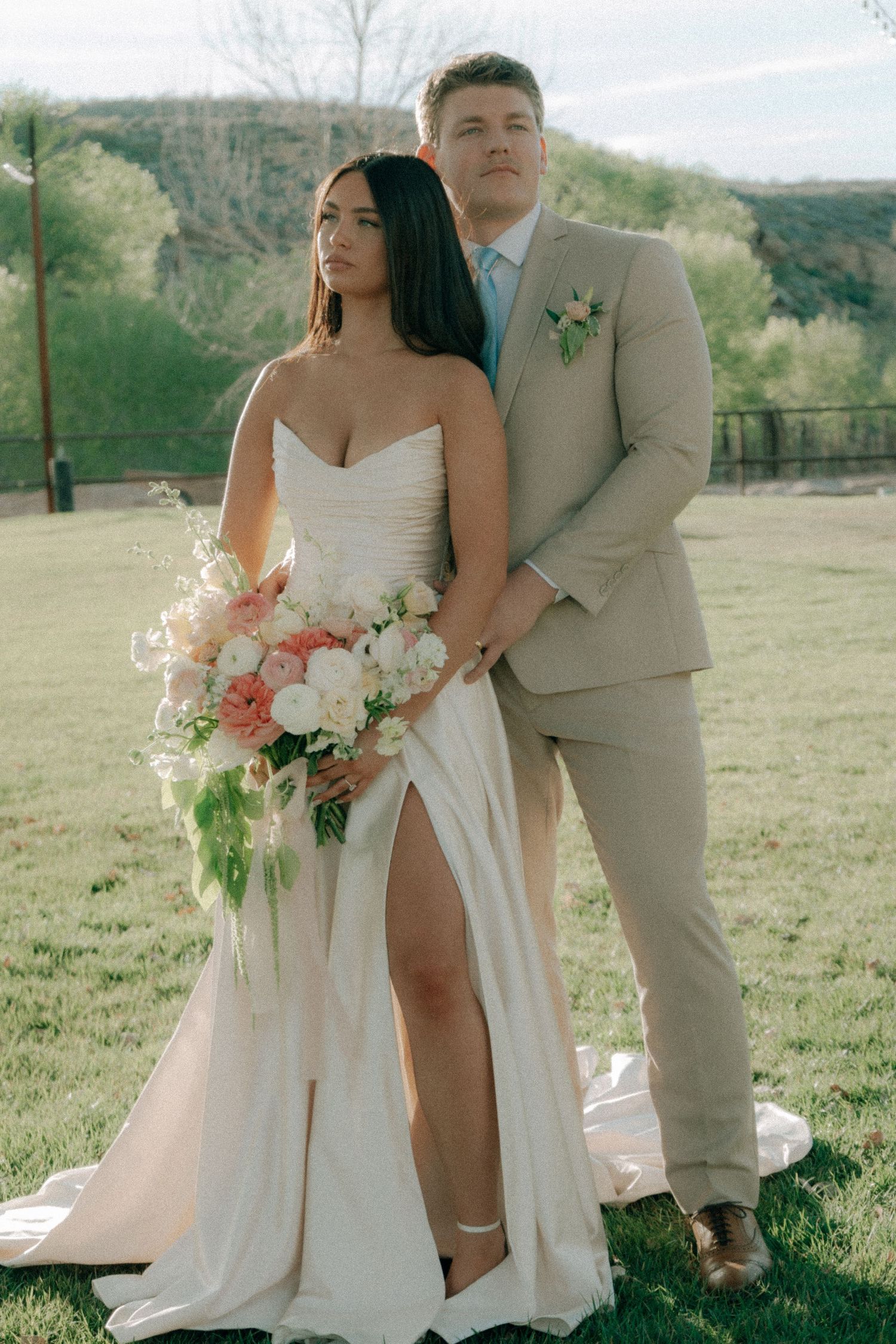 Bride in strapless white gown with slit holds pink floral bouquet beside groom in tan suit outdoors.