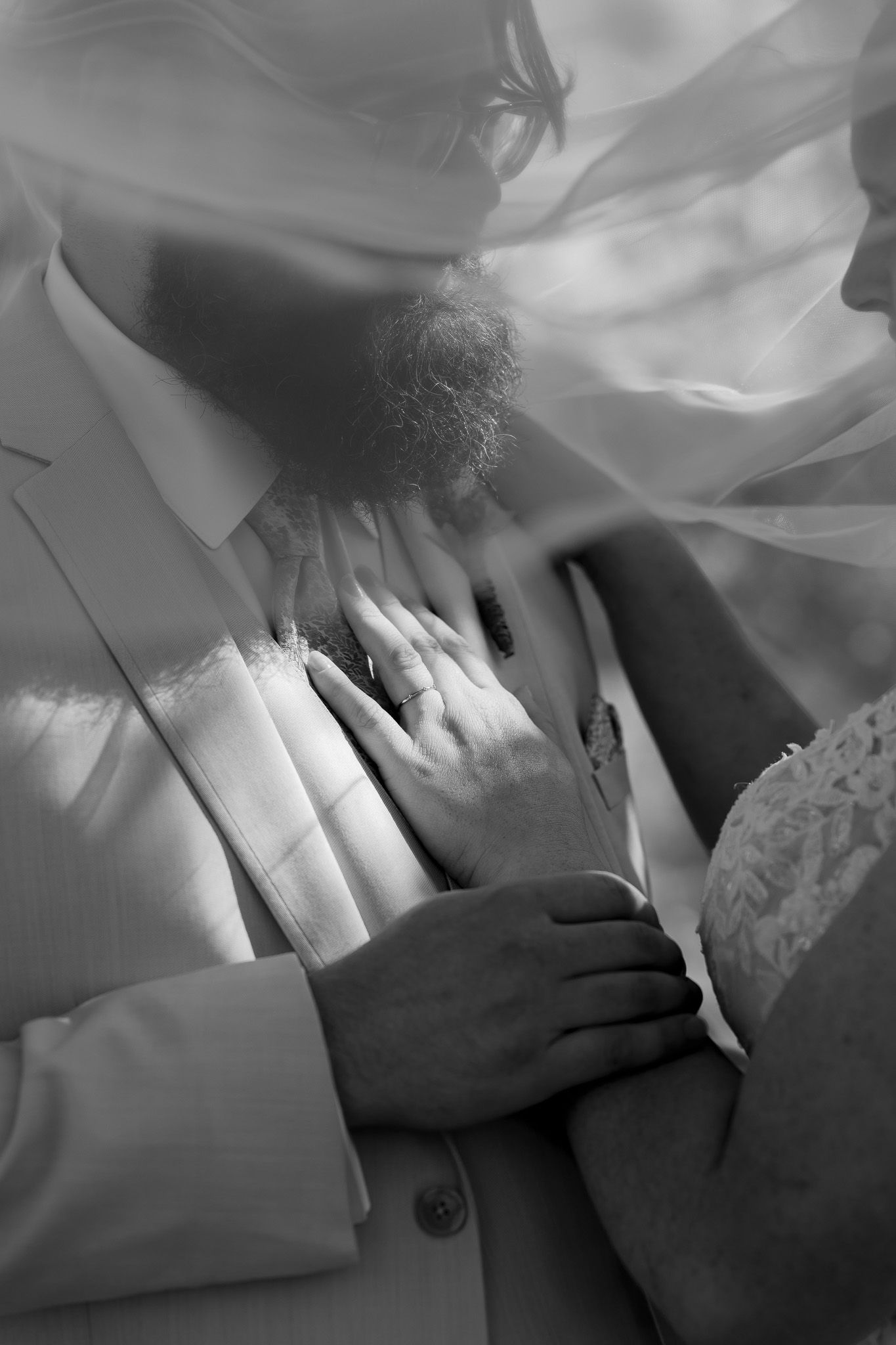 Black and white wedding photo of bride and groom embracing under veil, hands touching gently.