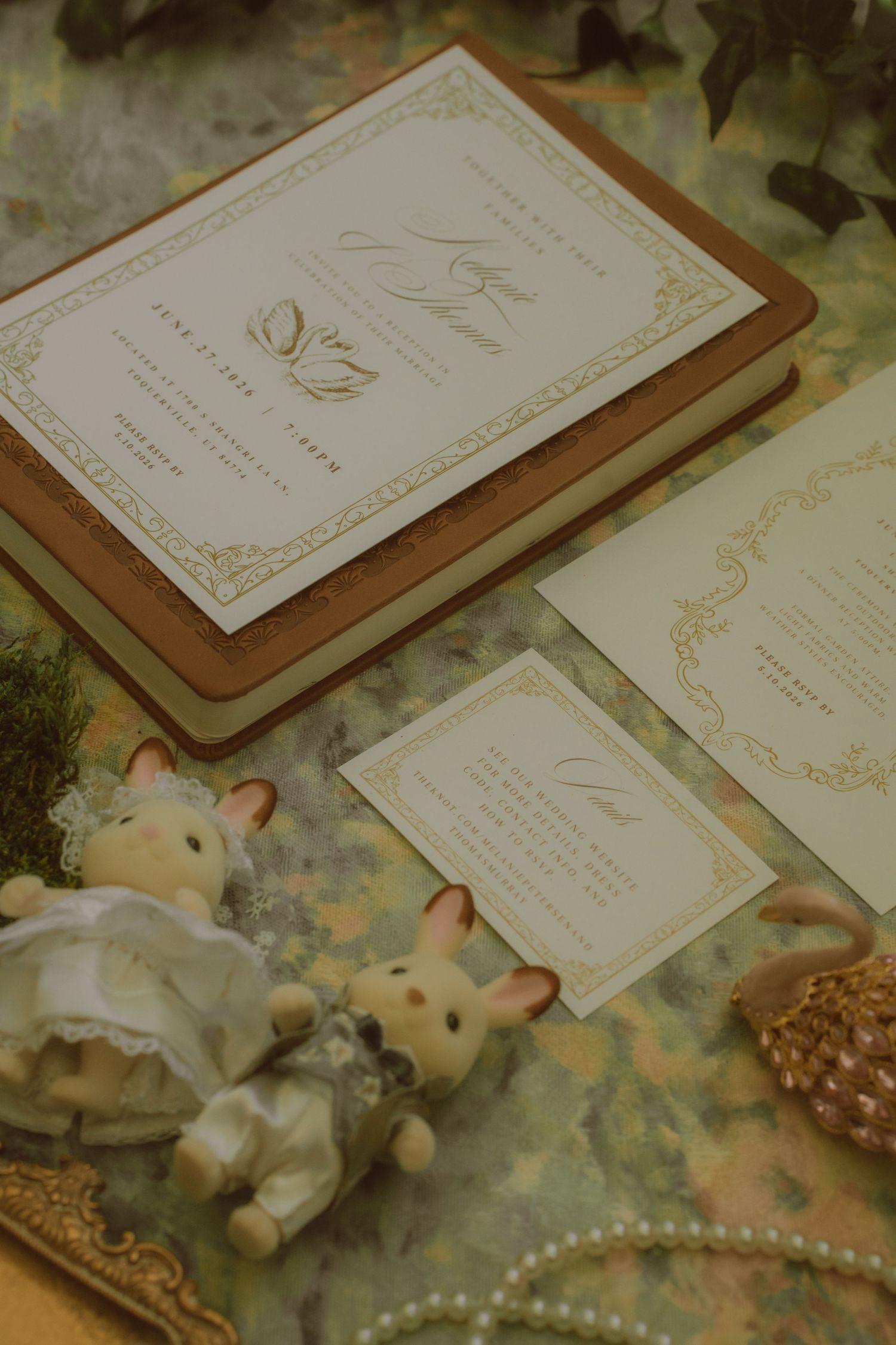 Elegant wedding invitation suite with gold foil details, displayed with Sylvanian Families figurines and pearl necklace on green surface.