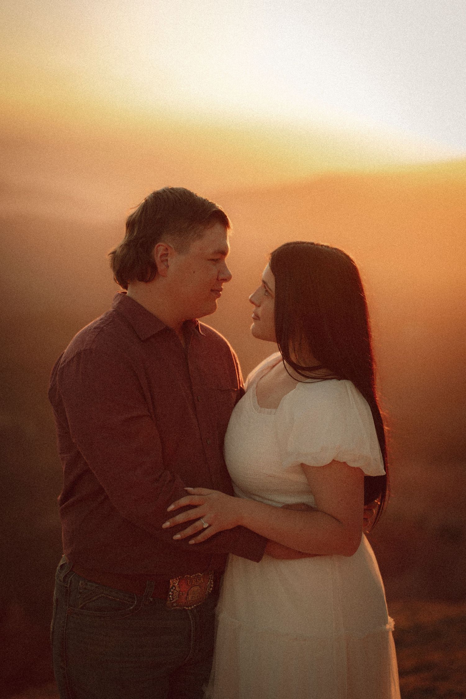 Couple embracing at golden hour sunset, man in red shirt and woman in white dress sharing a romantic moment outdoors.