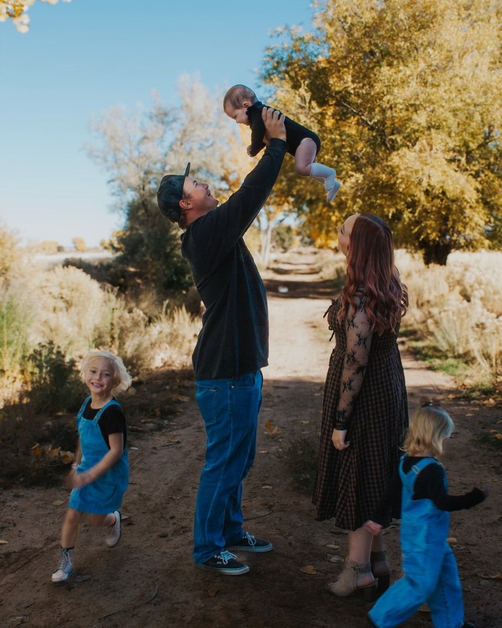 Family enjoying outdoor autumn day, father lifting baby in air while children play on dirt path surrounded by golden trees.