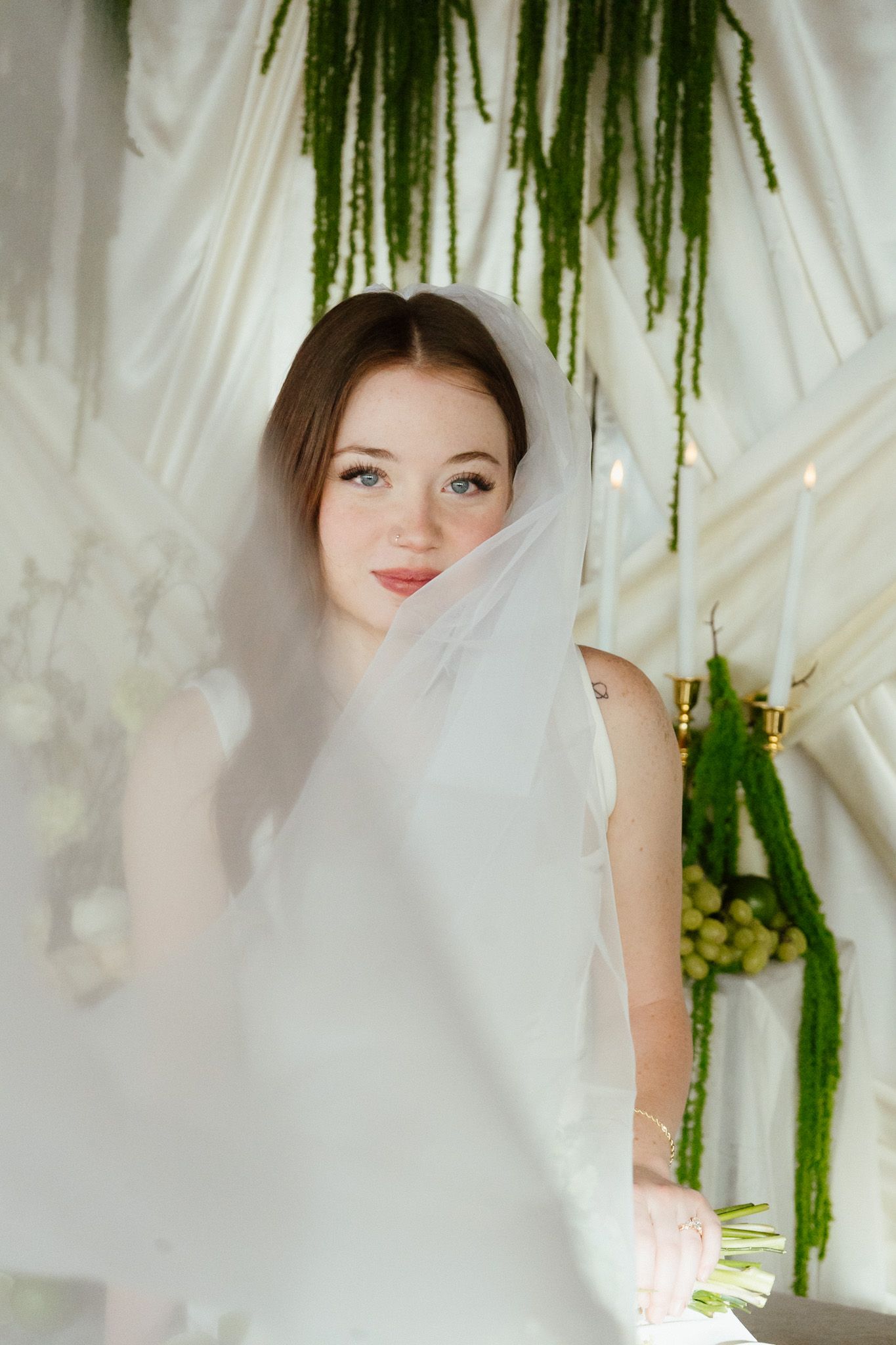 Beautiful bride with blue eyes wearing white veil poses in front of green floral wedding backdrop.