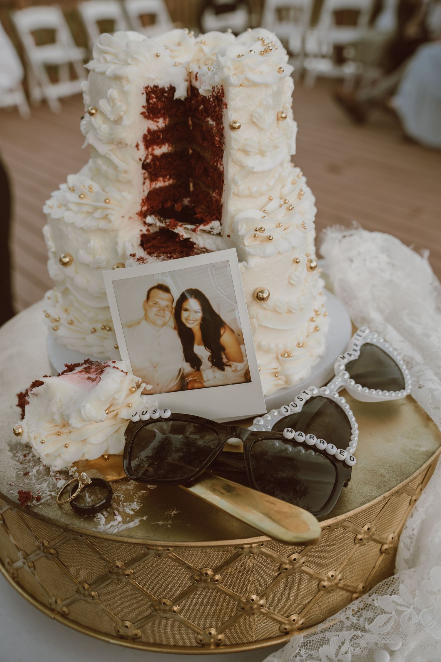 Red velvet wedding cake with white frosting, gold pearls, Polaroid photo, heart sunglasses on gold tray.