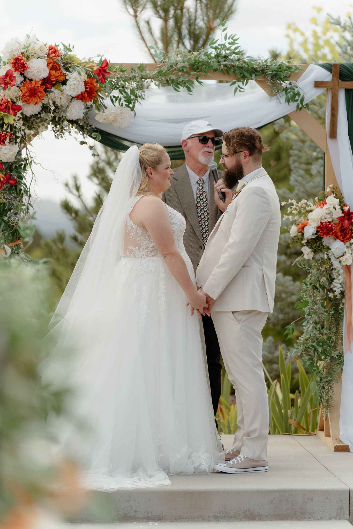 Bride and groom hold hands at outdoor wedding altar decorated with red and white floral arrangements and greenery.
