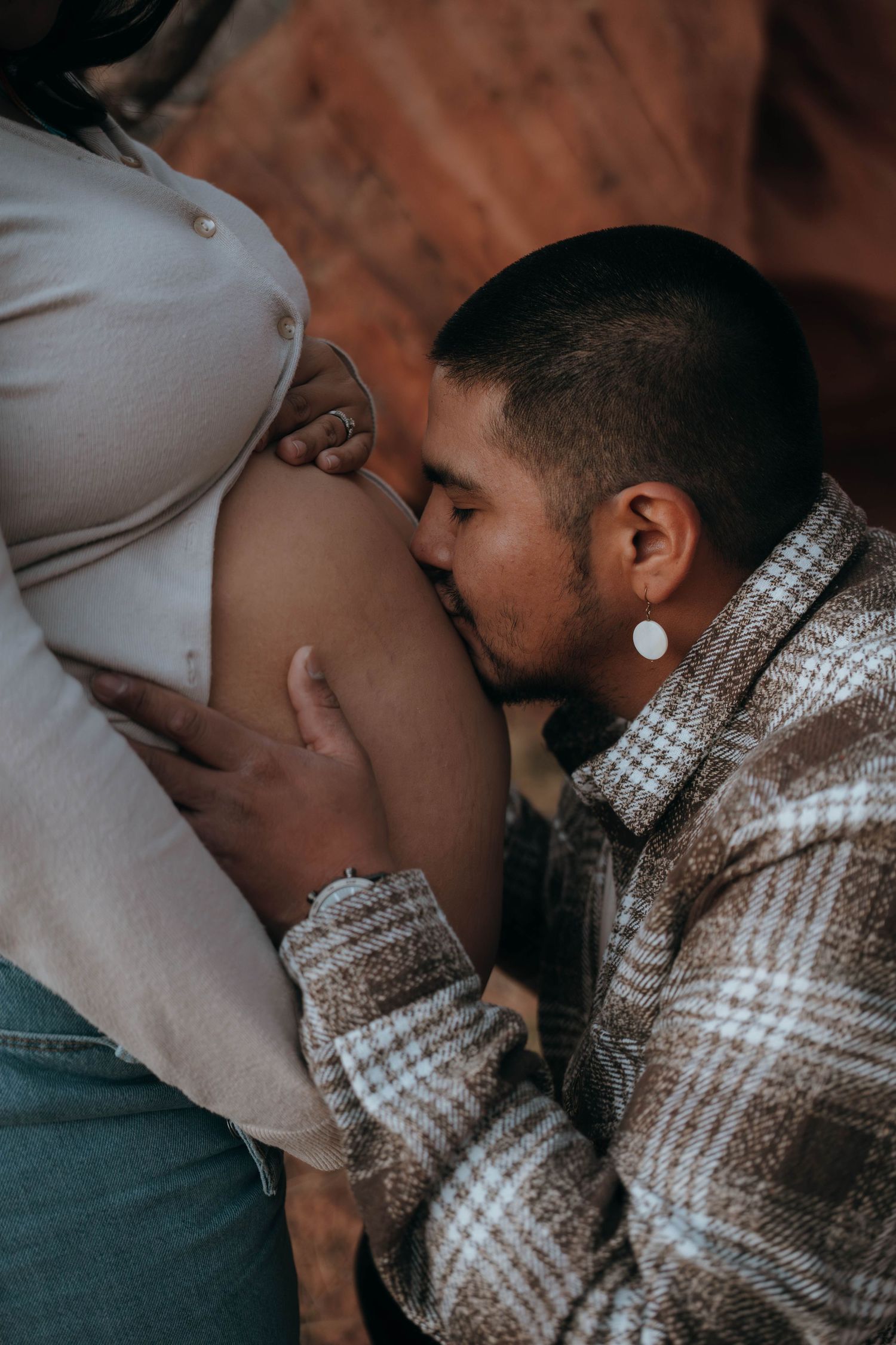 A man kisses a pregnant woman's belly during a maternity photoshoot, wearing a plaid flannel shirt with gauge earrings.