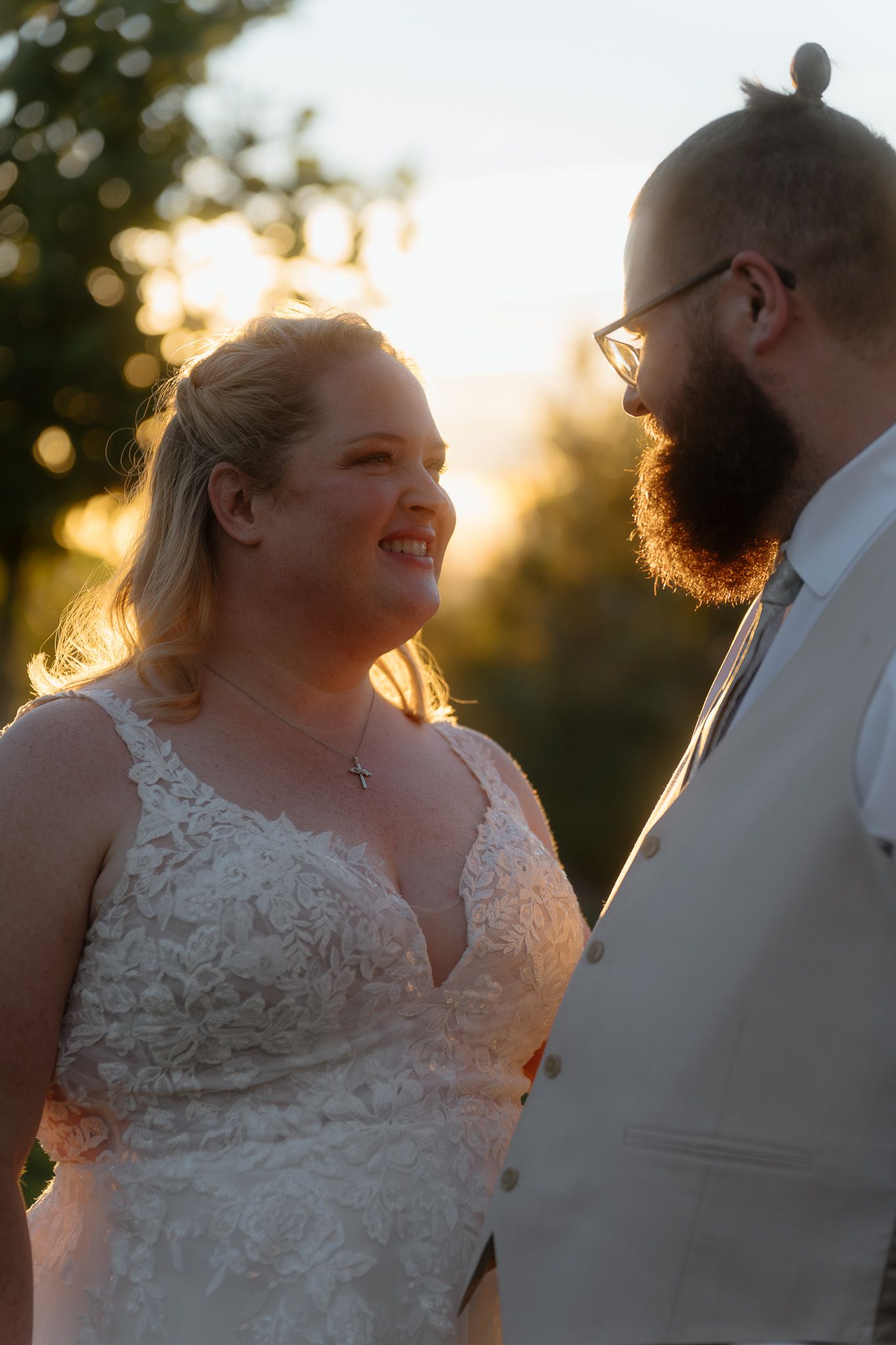 Bride in lace gown smiles at bearded groom in white suit during golden hour outdoor wedding portrait.
