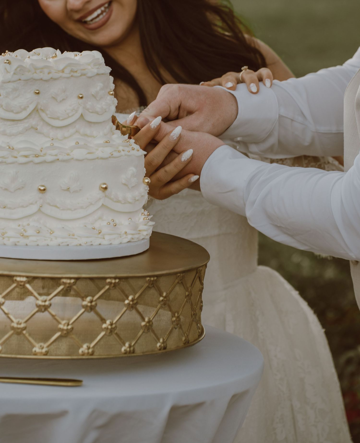 Couple cutting a two-tier white wedding cake with gold accents on a decorative gold stand outdoors.