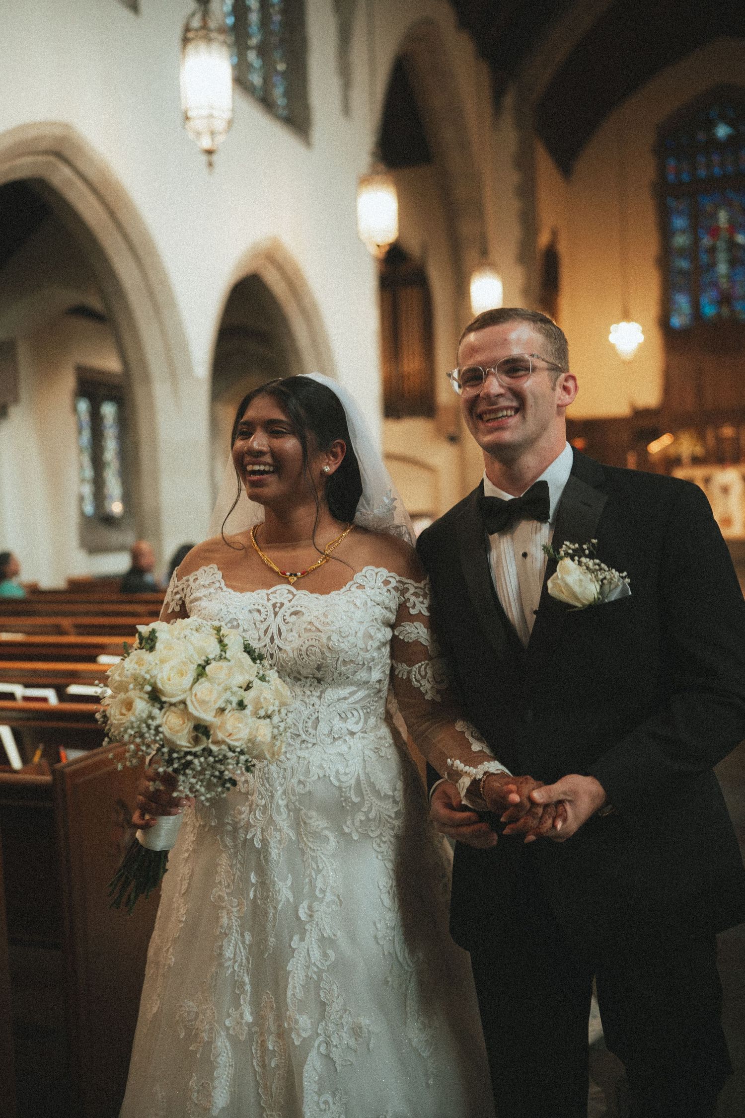 Joyful newlyweds walk down church aisle, bride in lace gown holding white bouquet, groom in black tuxedo.