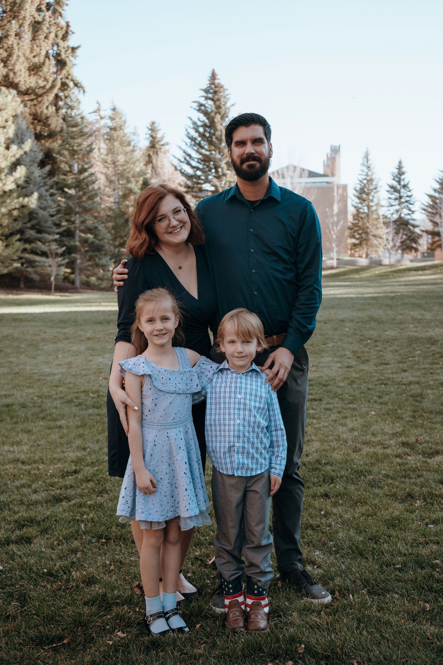 Family of four posing outdoors on green grass with tall evergreen trees in background during golden hour.
