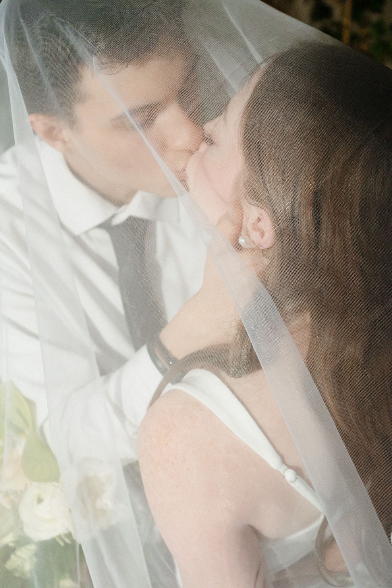 Bride and groom share a romantic kiss beneath a sheer white wedding veil outdoors.