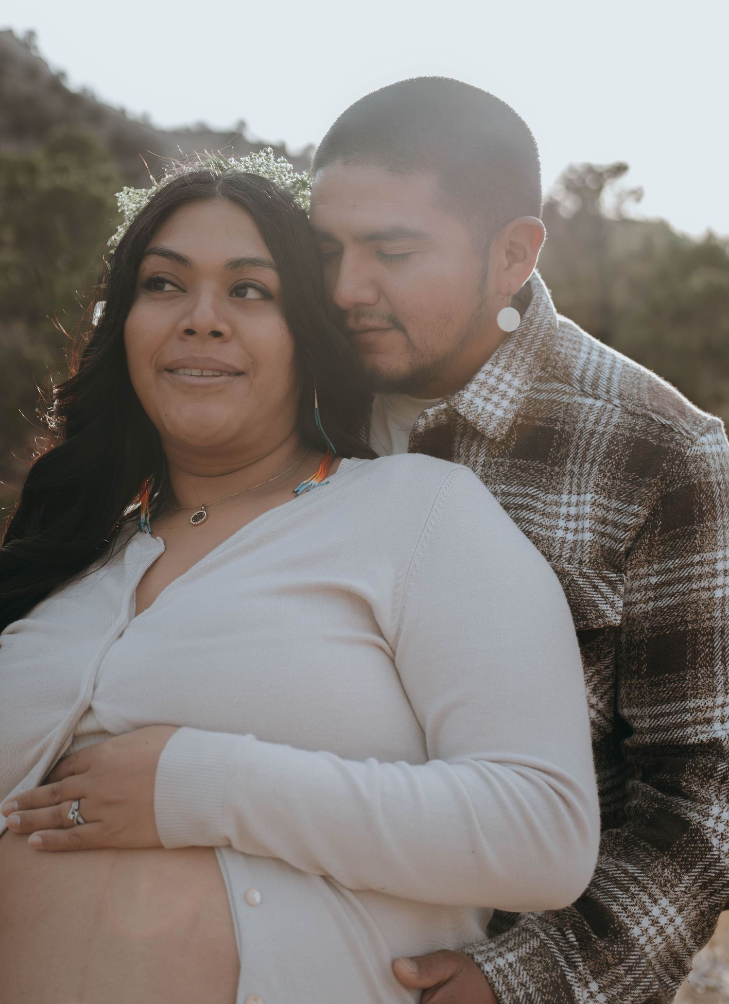 Couple sharing a tender maternity photo outdoors, woman in white dress with floral crown, man in plaid shirt embracing her baby bump.