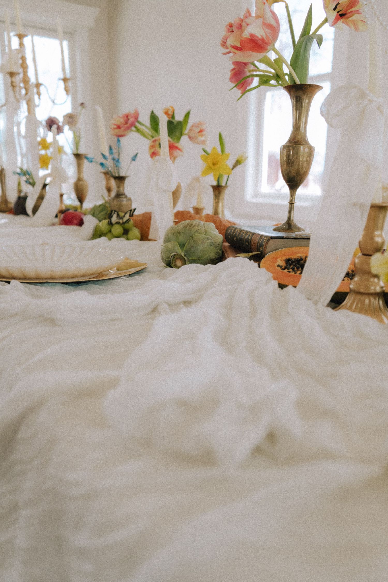Elegant spring tablescape with white linen, brass vases, tulips, fruits, and artichokes in a bright dining room.