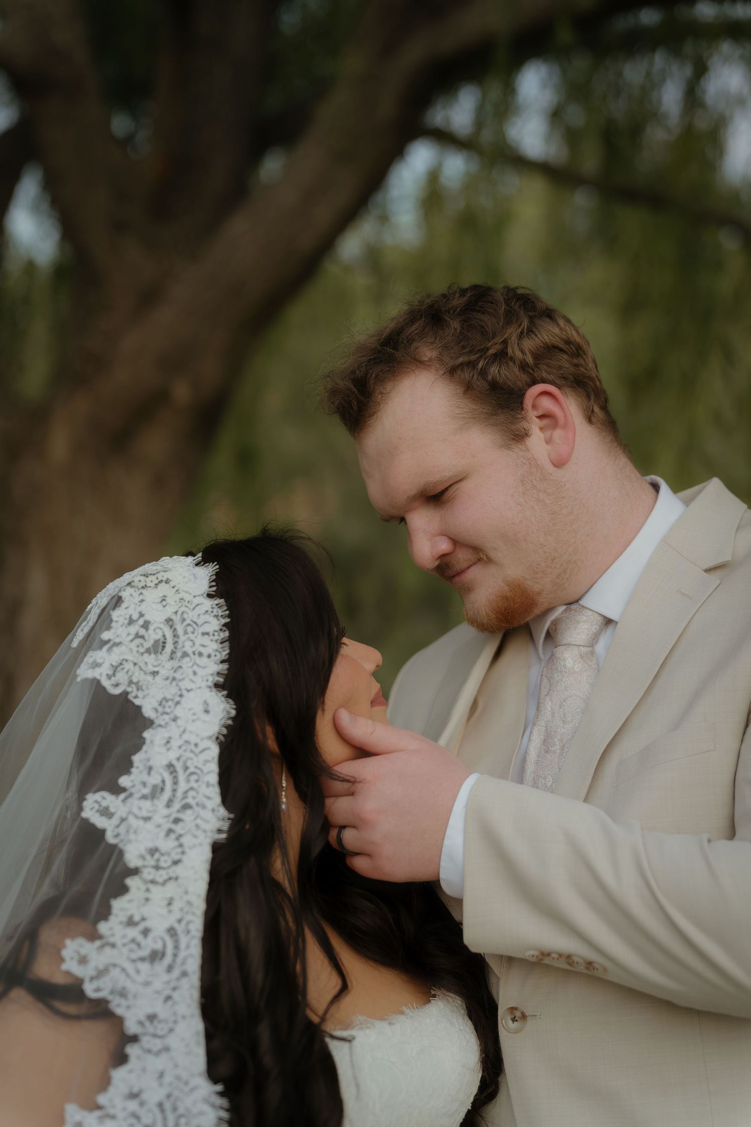 Groom in beige suit tenderly holds bride's face as she wears a lace veil, sharing an intimate moment outdoors.