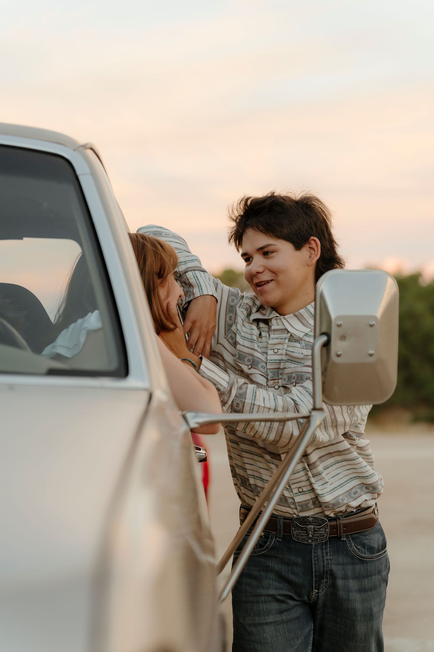 Two children leaning against a white truck at sunset, sharing a tender moment outdoors.