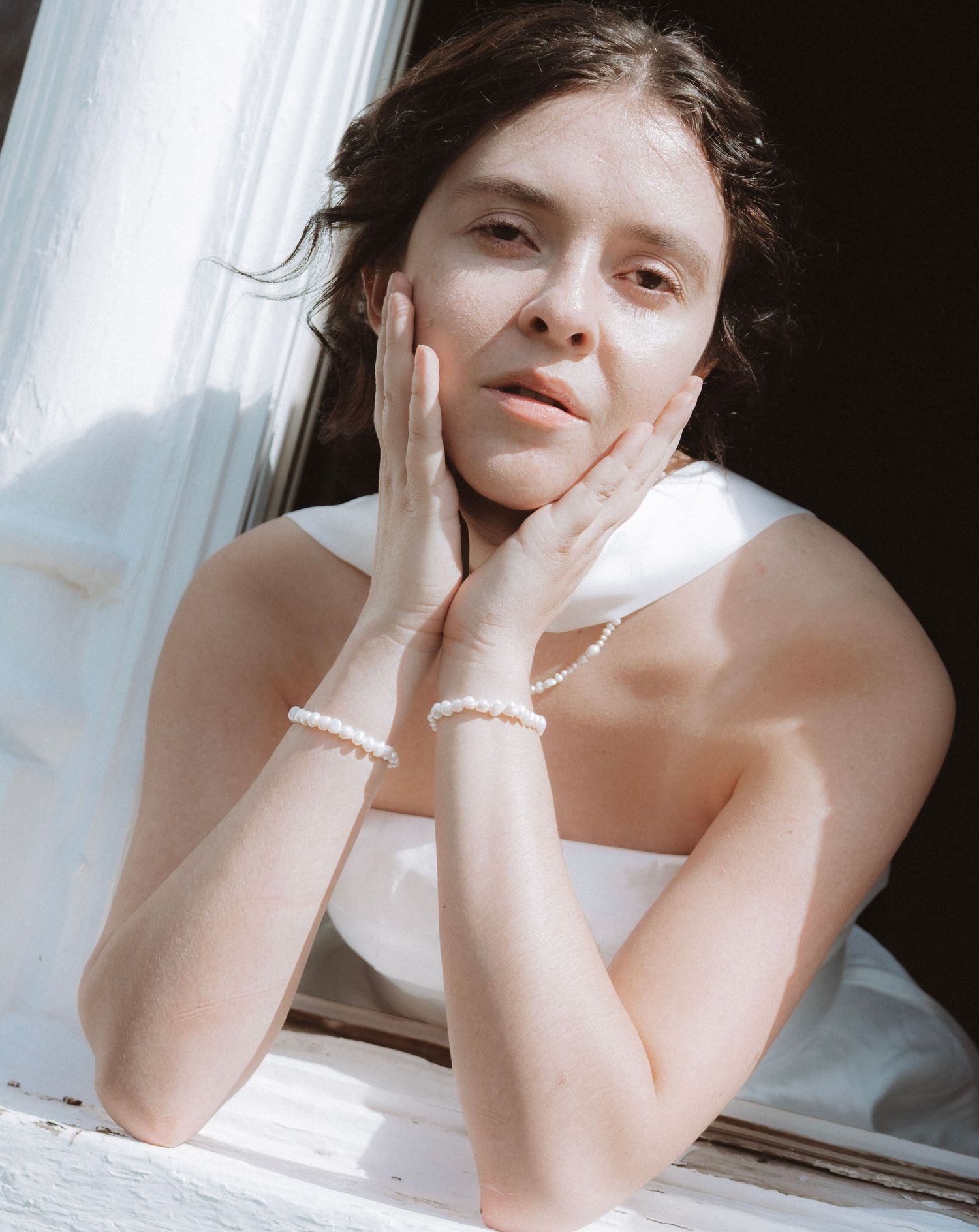 Radiant bride in white dress gazing out a sunlit window with pearl bracelets on her wrists.