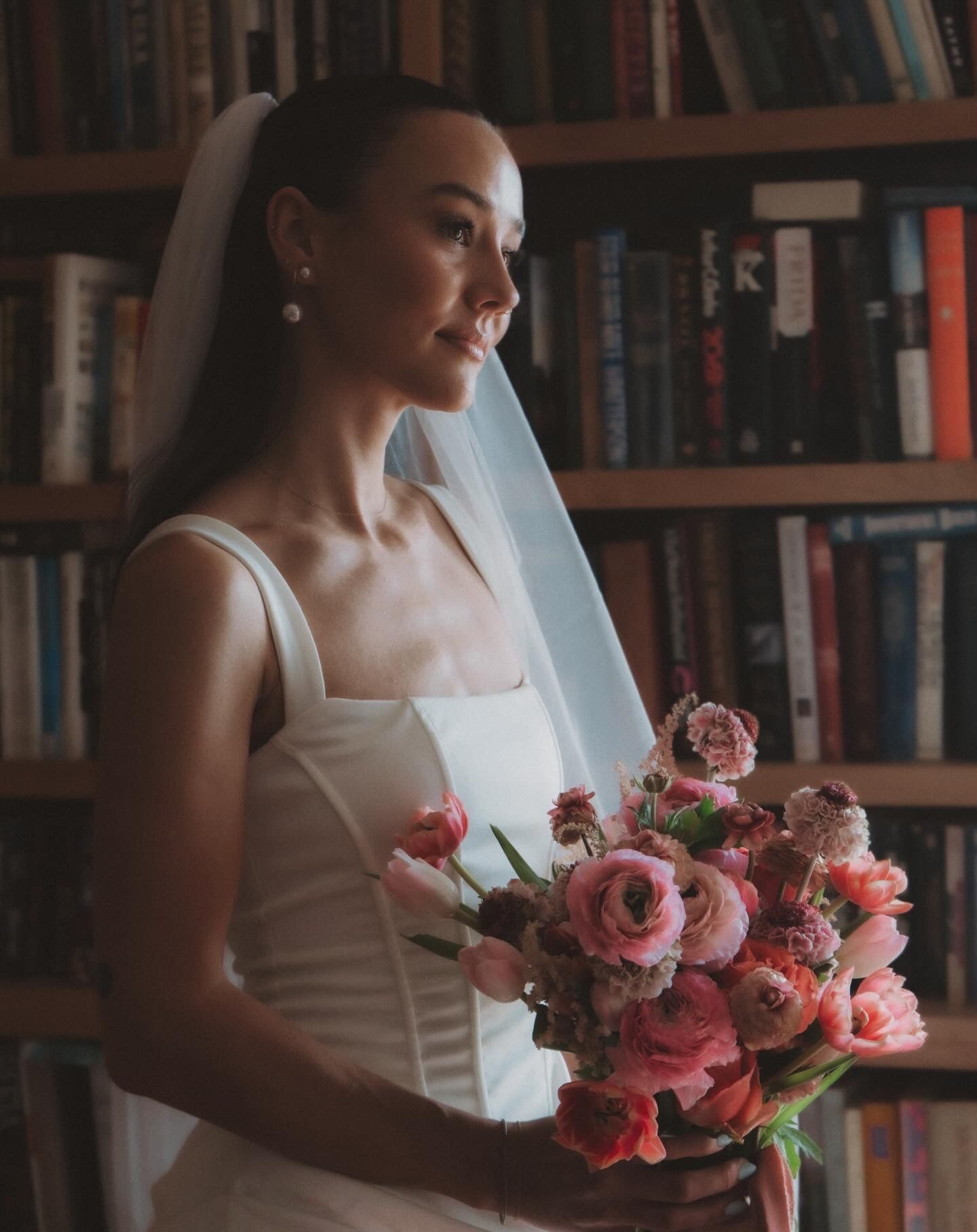 Bride in a sleek white gown holds a lush pink bouquet of ranunculus and tulips in a book-lined library.