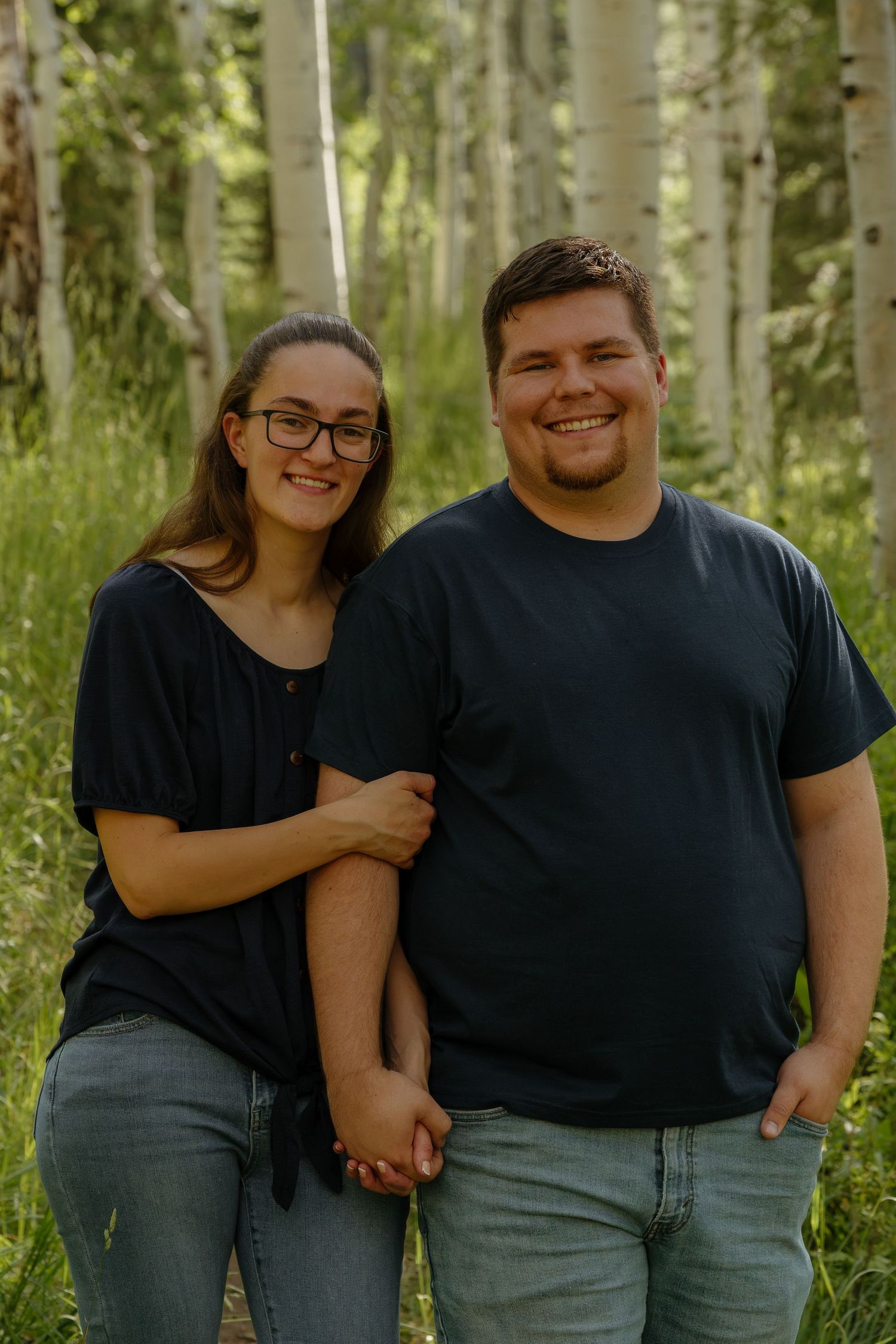 A smiling couple holds hands in a lush aspen forest, both wearing black tops and jeans.