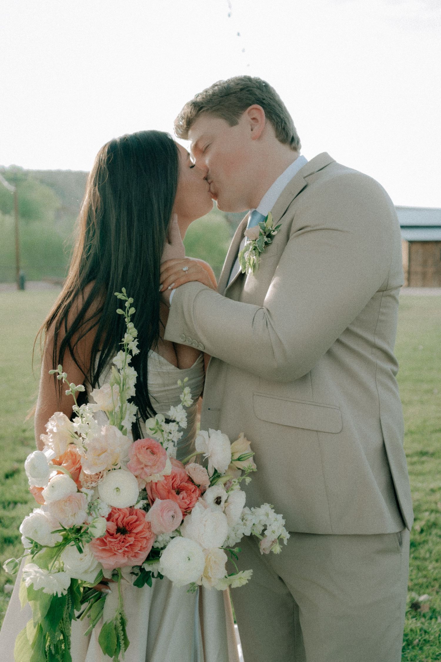 A bride and groom share a romantic kiss outdoors, she holds a lush pink and white floral bouquet, he wears a tan suit.