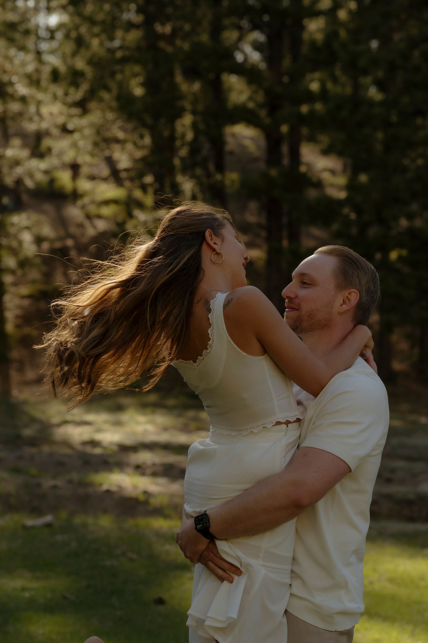 A man lifts a woman in a white dress outdoors, her long hair flowing, surrounded by lush green trees in warm sunlight.