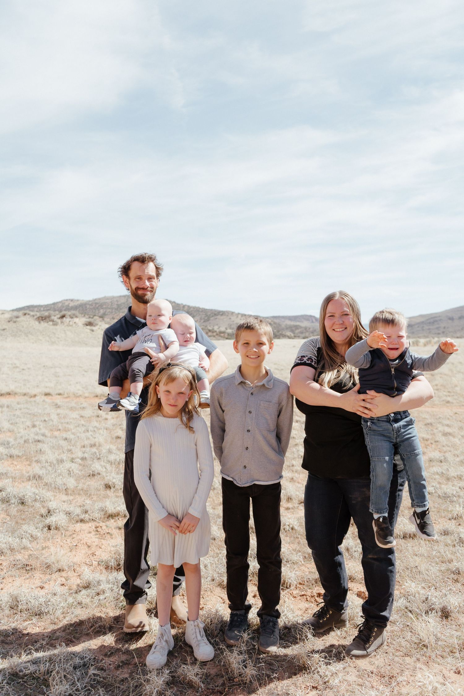 Family of six posing outdoors in a dry desert landscape, with parents holding young children under a cloudy sky.