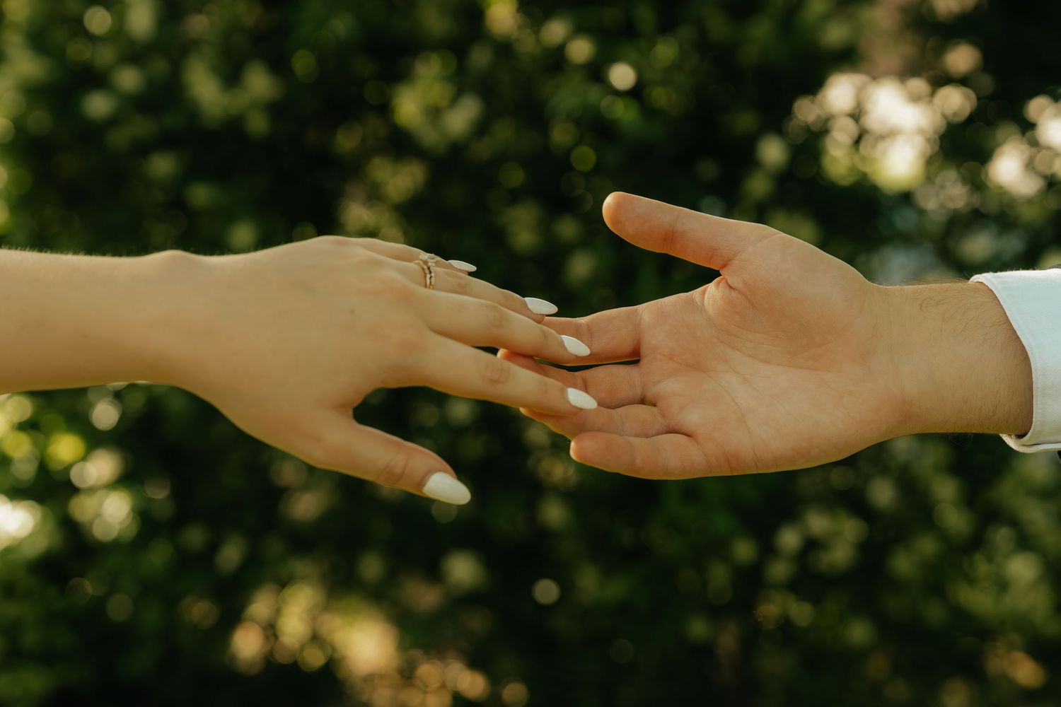 Two hands touching, woman wearing engagement ring with white manicure, green bokeh background.