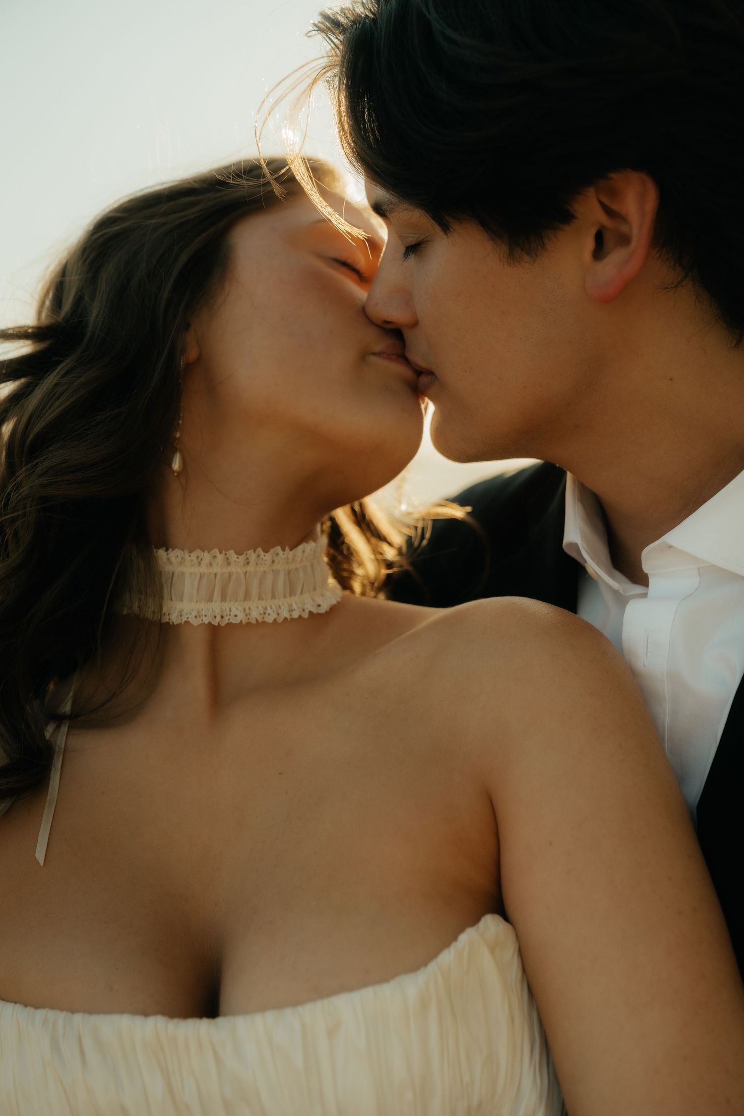 A bride and groom share a romantic kiss, with the bride wearing a white dress and lace choker necklace.