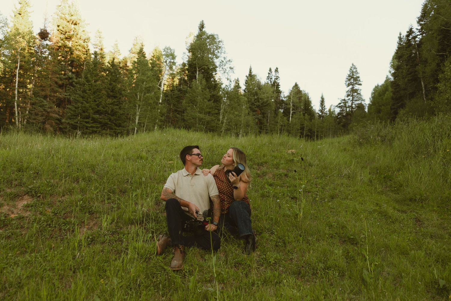 Couple sitting together in a lush green meadow surrounded by tall pine trees, sharing a romantic outdoor moment.