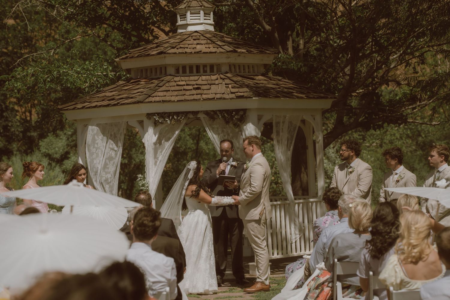 Outdoor wedding ceremony under a white gazebo surrounded by lush greenery with bride, groom, and officiant.
