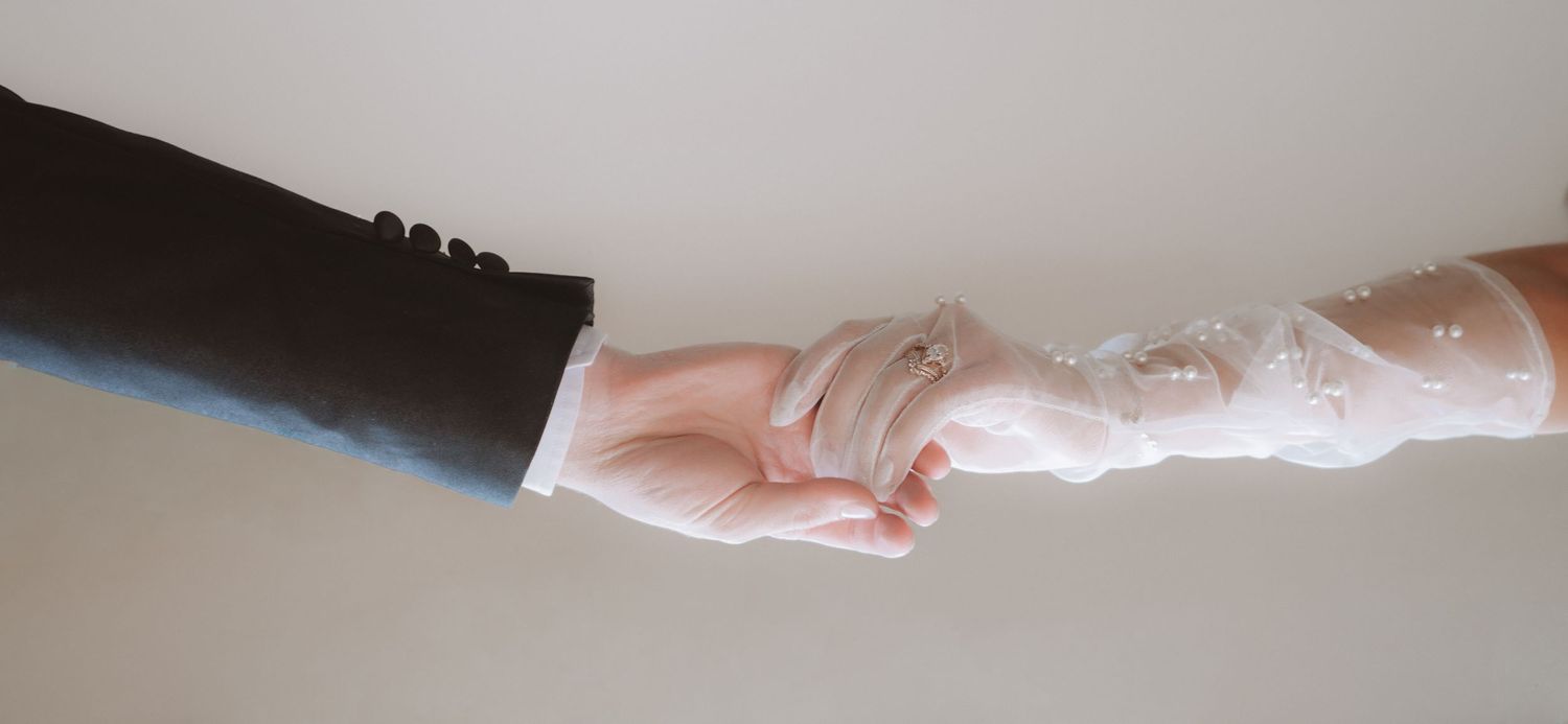 Bride in lace glove and groom in black suit hold hands in elegant wedding moment.